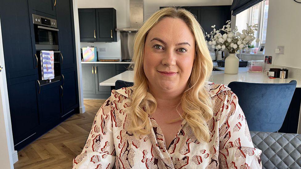 A woman with blonde shoulder-length hair wearing a light-coloured patterned shirt. She is sitting in her kitchen.