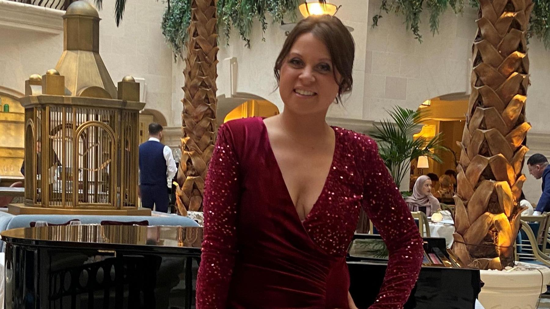 A woman with dark brown hair pinned up. She is wearing a long burgundy dress and behind her is a grand piano, palm tress and table and chairs. 