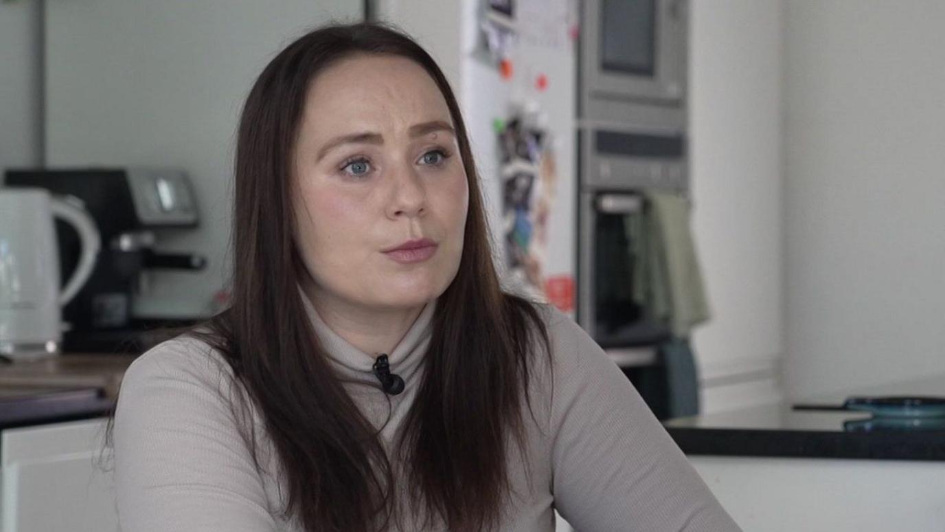 Rosanna Brown is sitting in a kitchen with the fridge and countertop out of focus behind her. She has long brown hair over her shoulders and is wearing a beige polo neck top. 'I made a choice - I didn't naturally lose my baby'