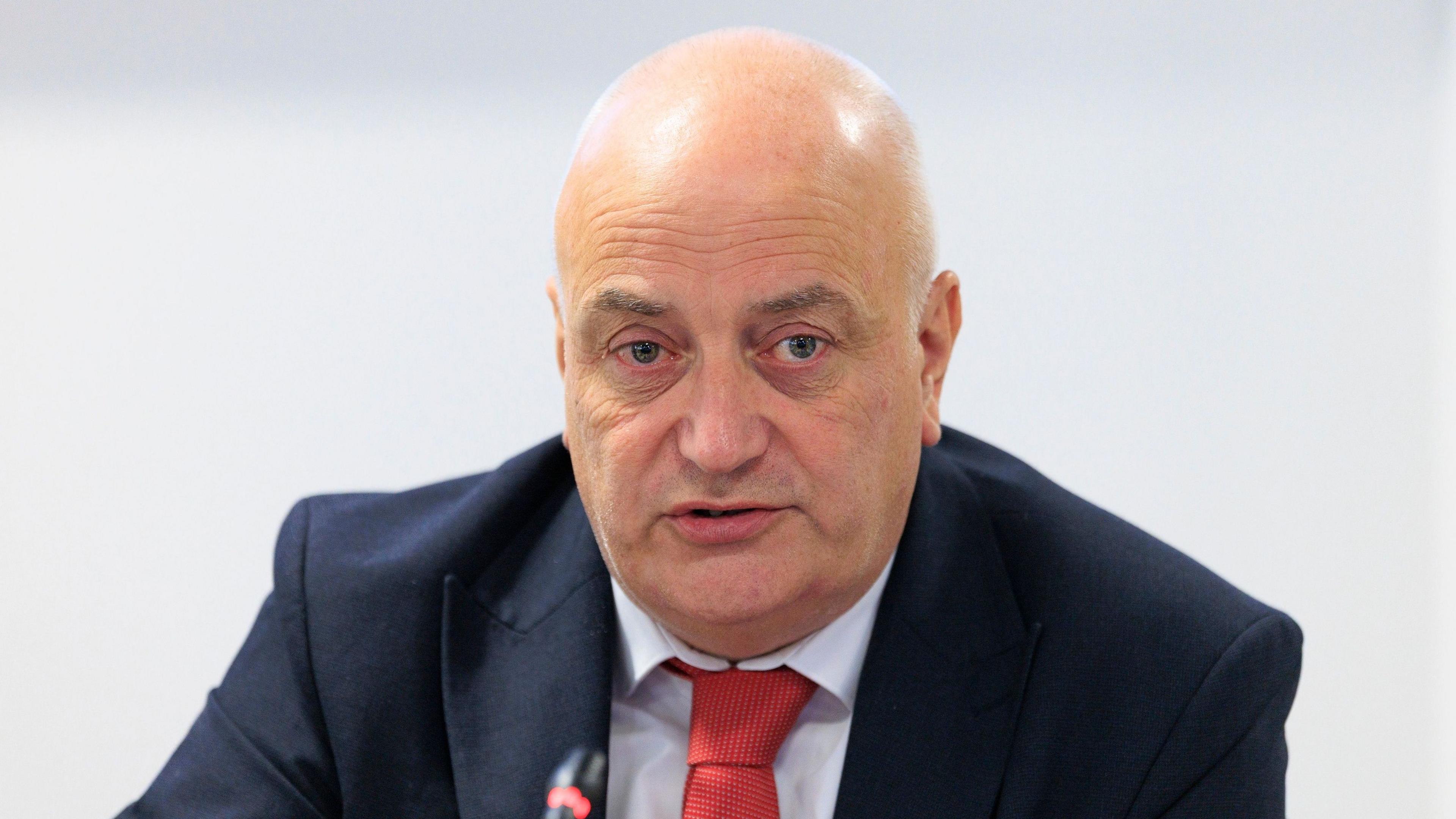 Trevor Clarke, a man with a shaved head, speaks during a Policing Board meeting in September 2026. He is wearing a navy suit jacket, a white shirt and a red tie.