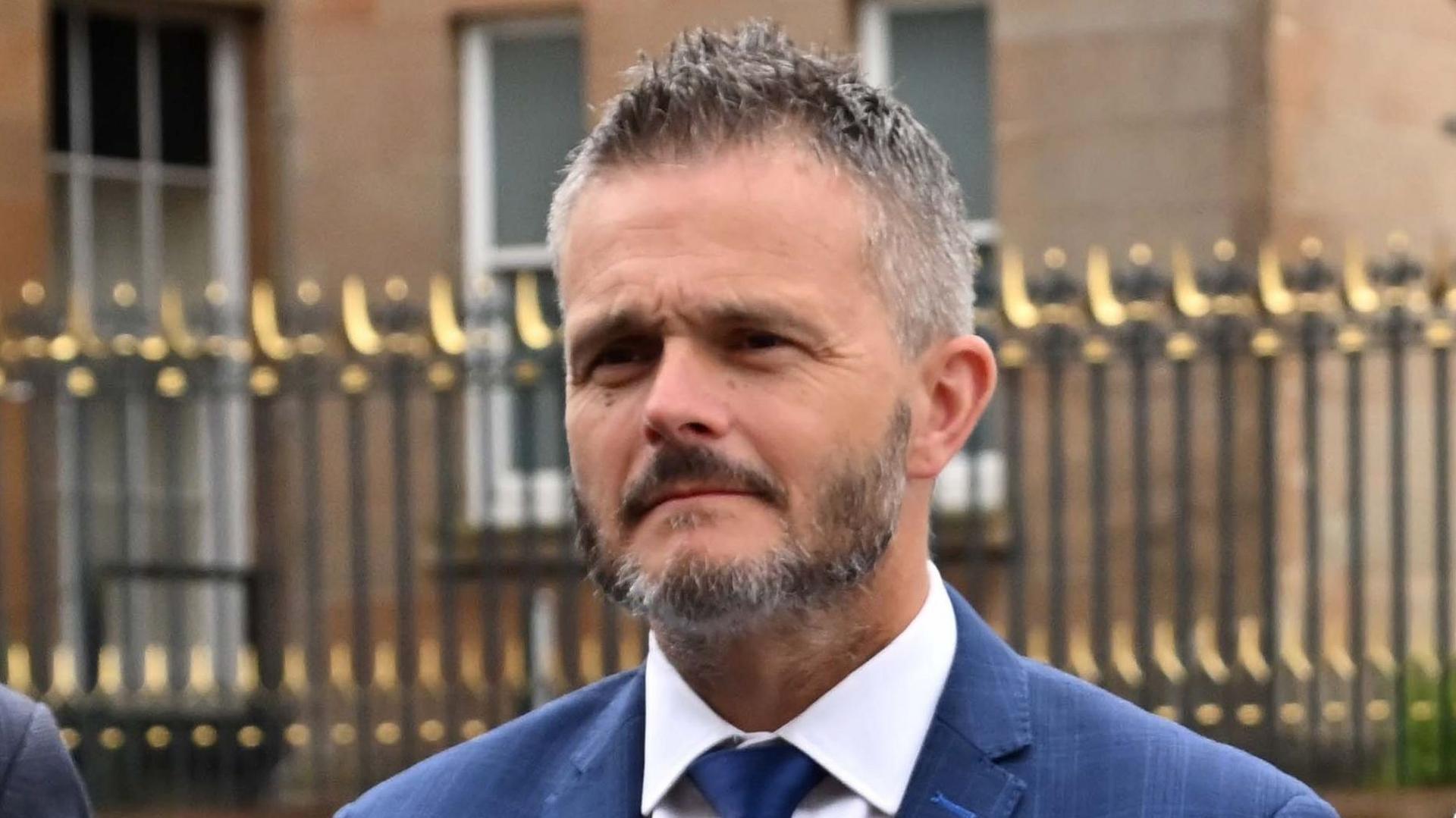 Robbie Butler, a man with short grey hair and a matching beard, standing outside Hillsborough Castle. He is wearing a blue suit, white shirt and blue tie. There are black and gold metal railings around the Georgian building behind him.