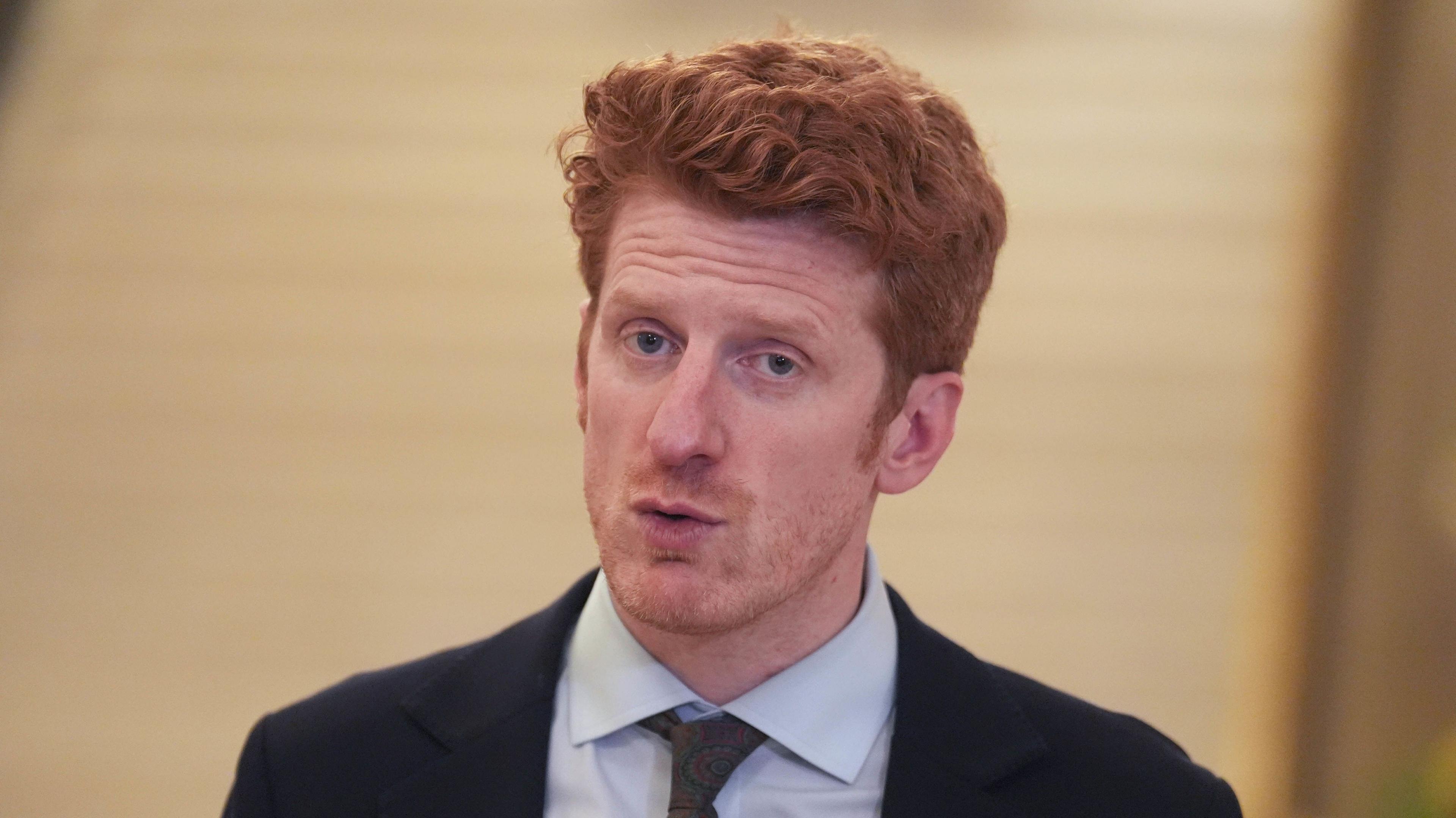 A man with short ginger curly hair wearing a black blazer, white shirt and dark tie.