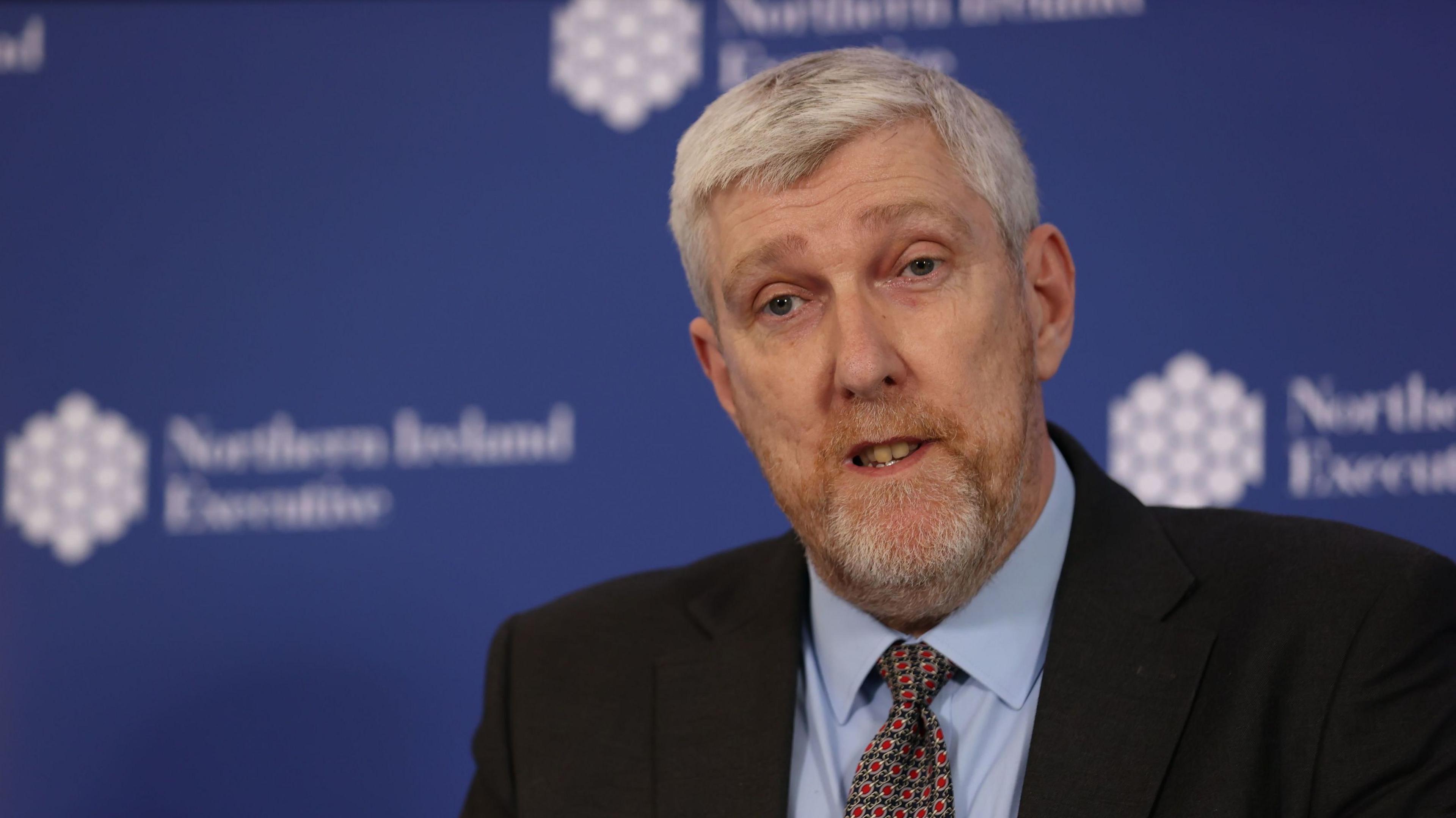 John O'Dowd. He has short white hair and beard, wearing a black blazer, light blue shirt and tie. Blue NI executive branding is behind him.
