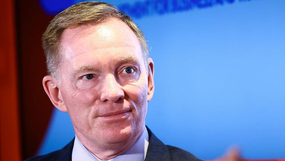A head-and-shoulders image of Sir Chris Bryant, who has short, fair hair and wears a dark navy suit jacket over a light blue shirt. He stands in front of a blue screen and is looking to his left.