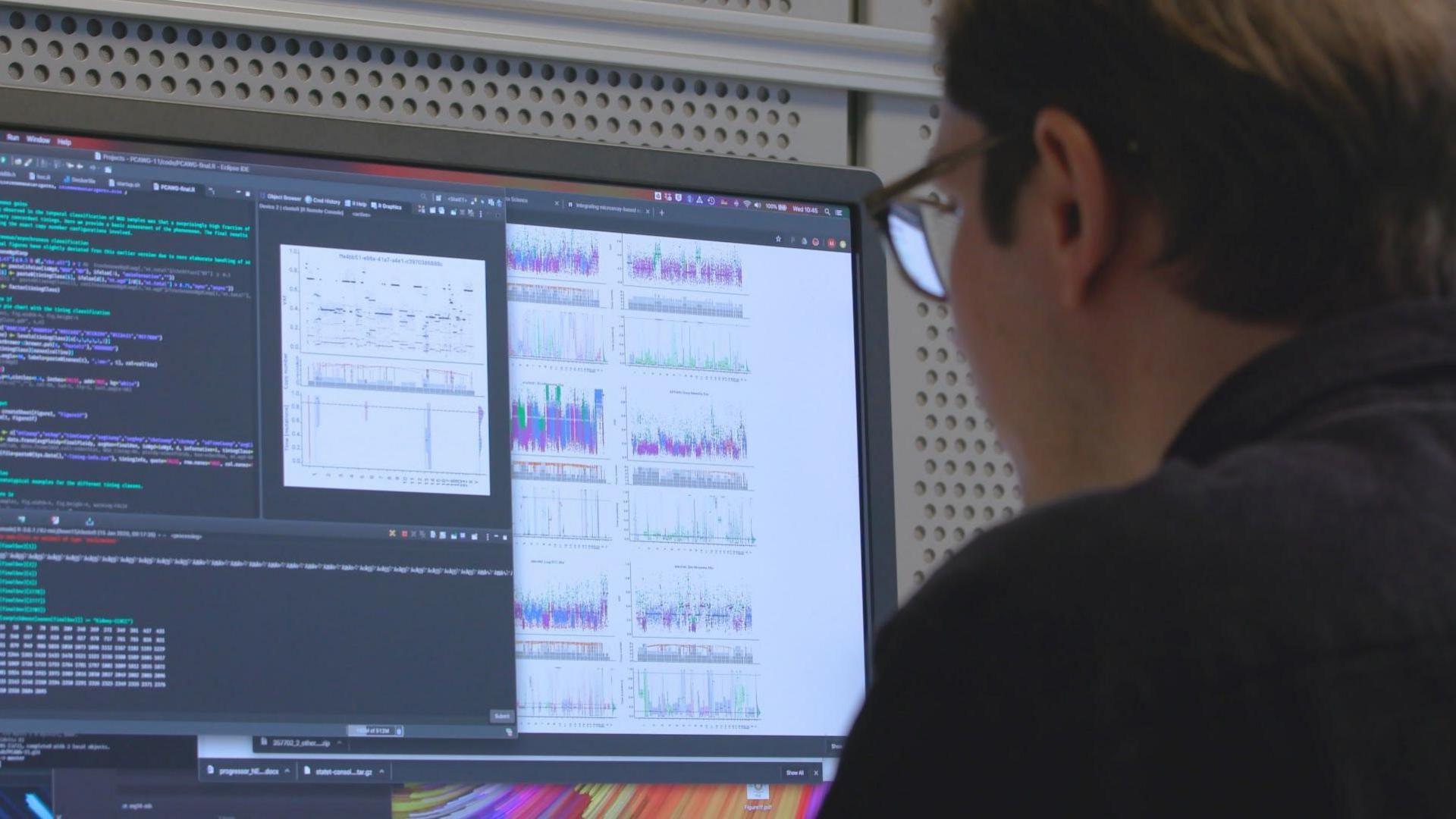 The back of a dark-haired man's head and shoulders as he looks at a computer screen. You can see lines of computer code and multicoloured graphs being produced although their meaning is unclear form the image. 