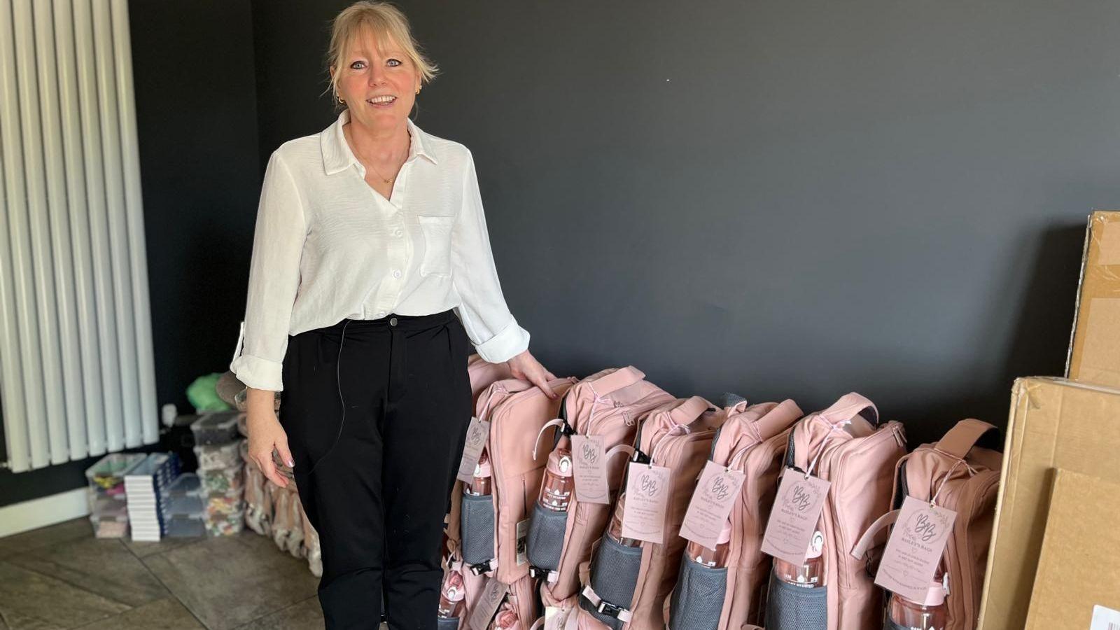Maria Bailey is standing in front of a row of pink matching backpacks with water bottles in the side pocket and a pink label on each. She is wearing black trousers and a white shirt