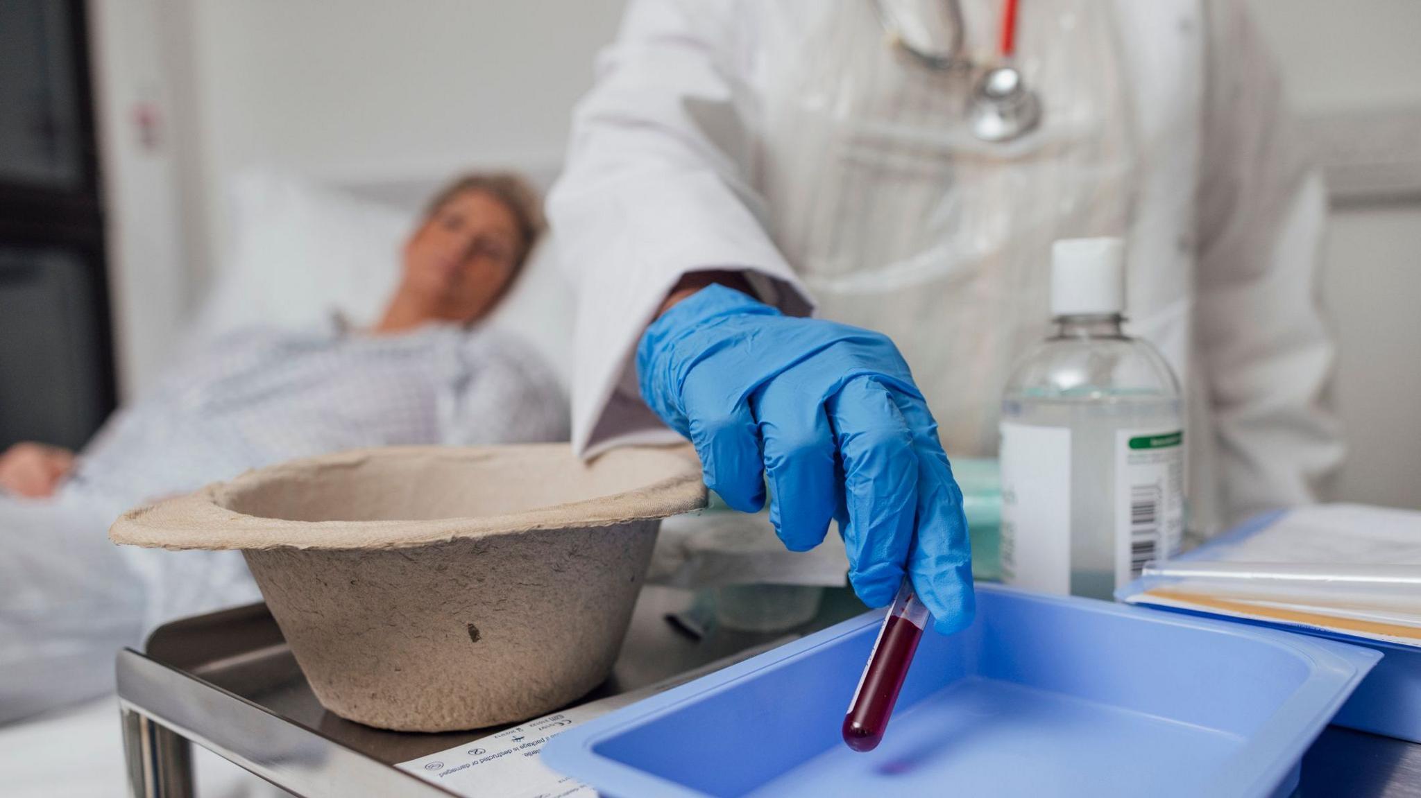A close up of a nurse's hand taking a blood sample from a woman who is undergoing treatment for cancer. The nurse is wearing a blue plastic glove and holding a test tube half filled with blood.