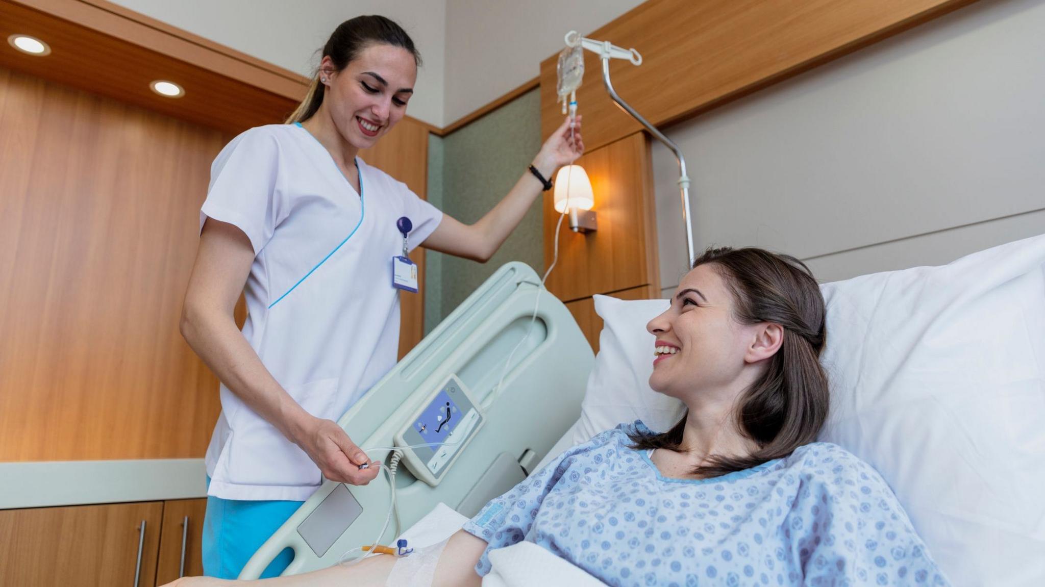A nurse hold's up a patients cannula taking a blood sample from a woman in a hospital bed who is undergoing treatment for cancer. The nurse is wearing a white tunic and bluie trousers - the patient has long brown hair and wears a blue surgical gown