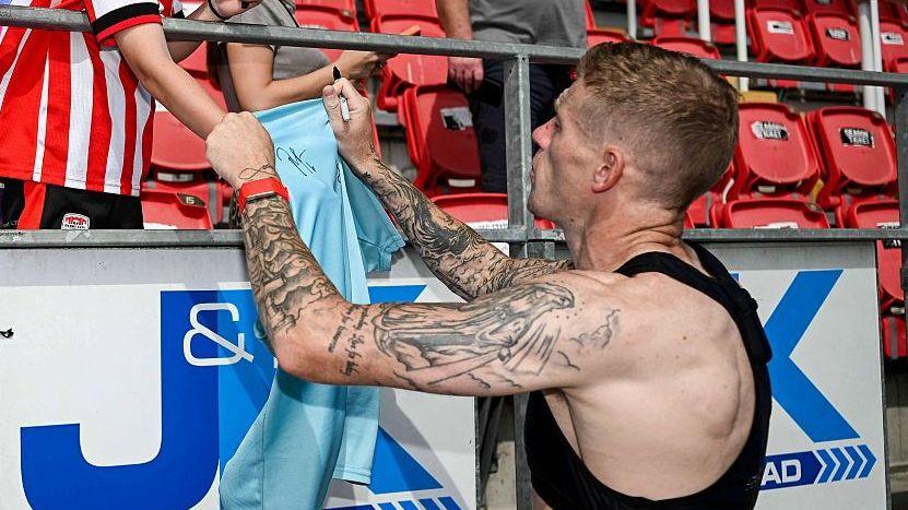 James McClean is pictured in a black vest signing a Derry City shirt for a young fan.