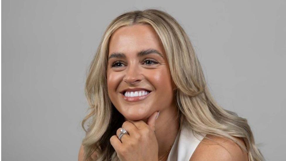 Shows a woman with blond hair smiling. She is wearing a wedding ring and a cream top