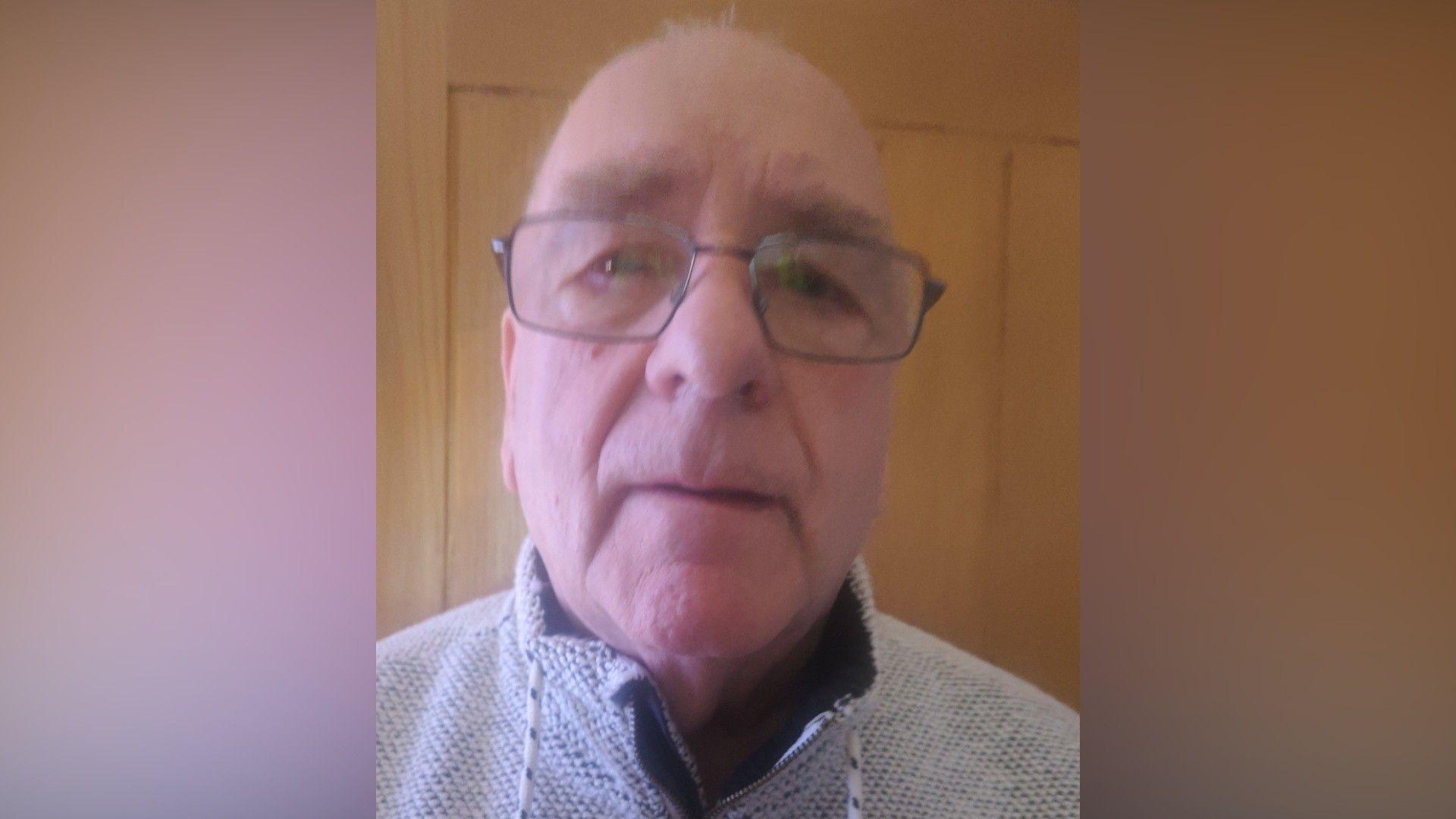 An older bald man who is wearing glasses and a grey woollen top with a high collar.