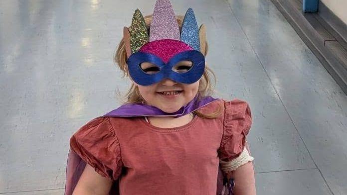 A young girl walking along a corridor dressed as a superhero. She is wearing a blue mask with a crown and a cape. She is looking at the camera and smiling.