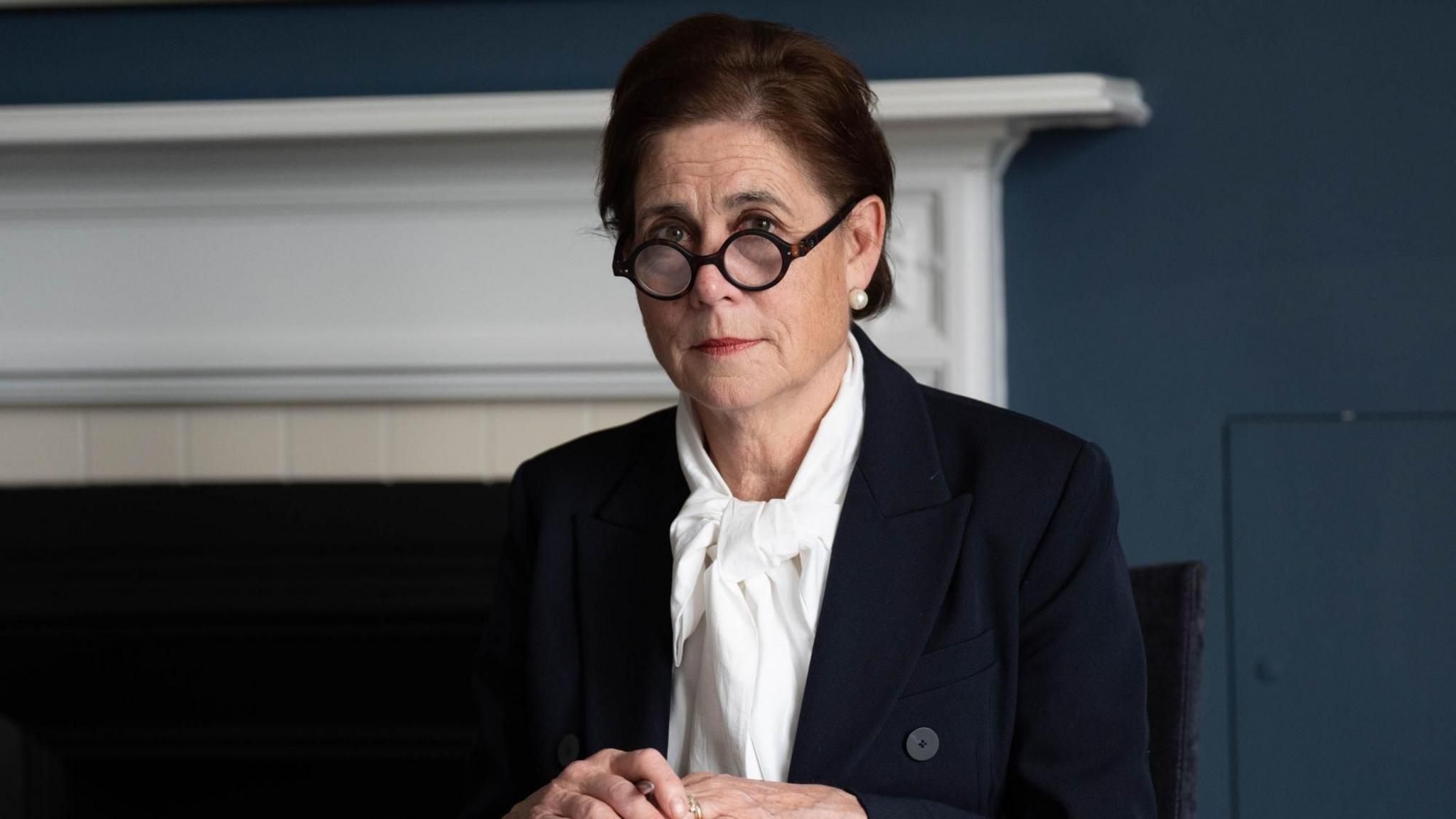 Baroness Kate Lampard sits at a table. She is wearing a black jacket and a white blouse with a tie at the neck. She has round framed tortoiseshell glasses and looks at the camera, sitting in front of the fireplace.