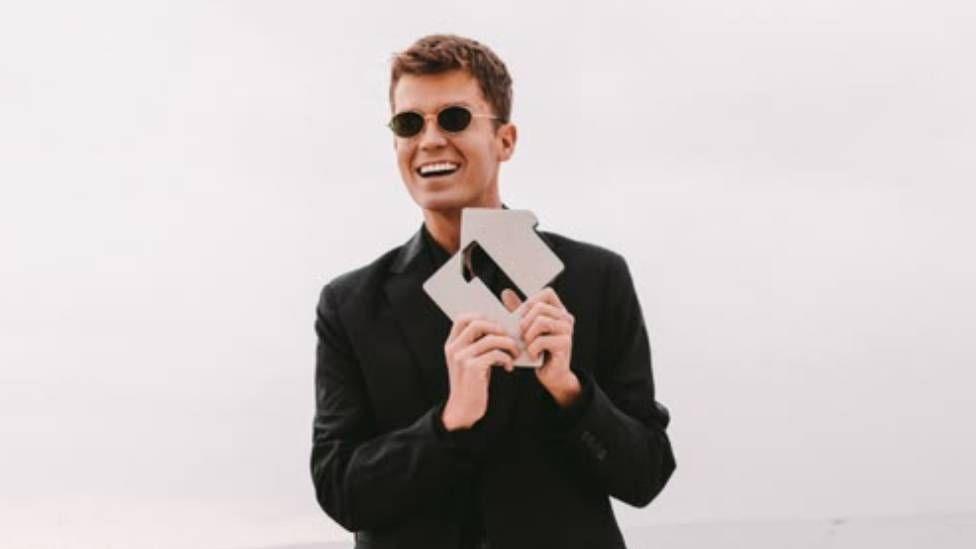A man is wearing glasses and a black suit outfit, holding an award, on a sandy beach in front of the sea.