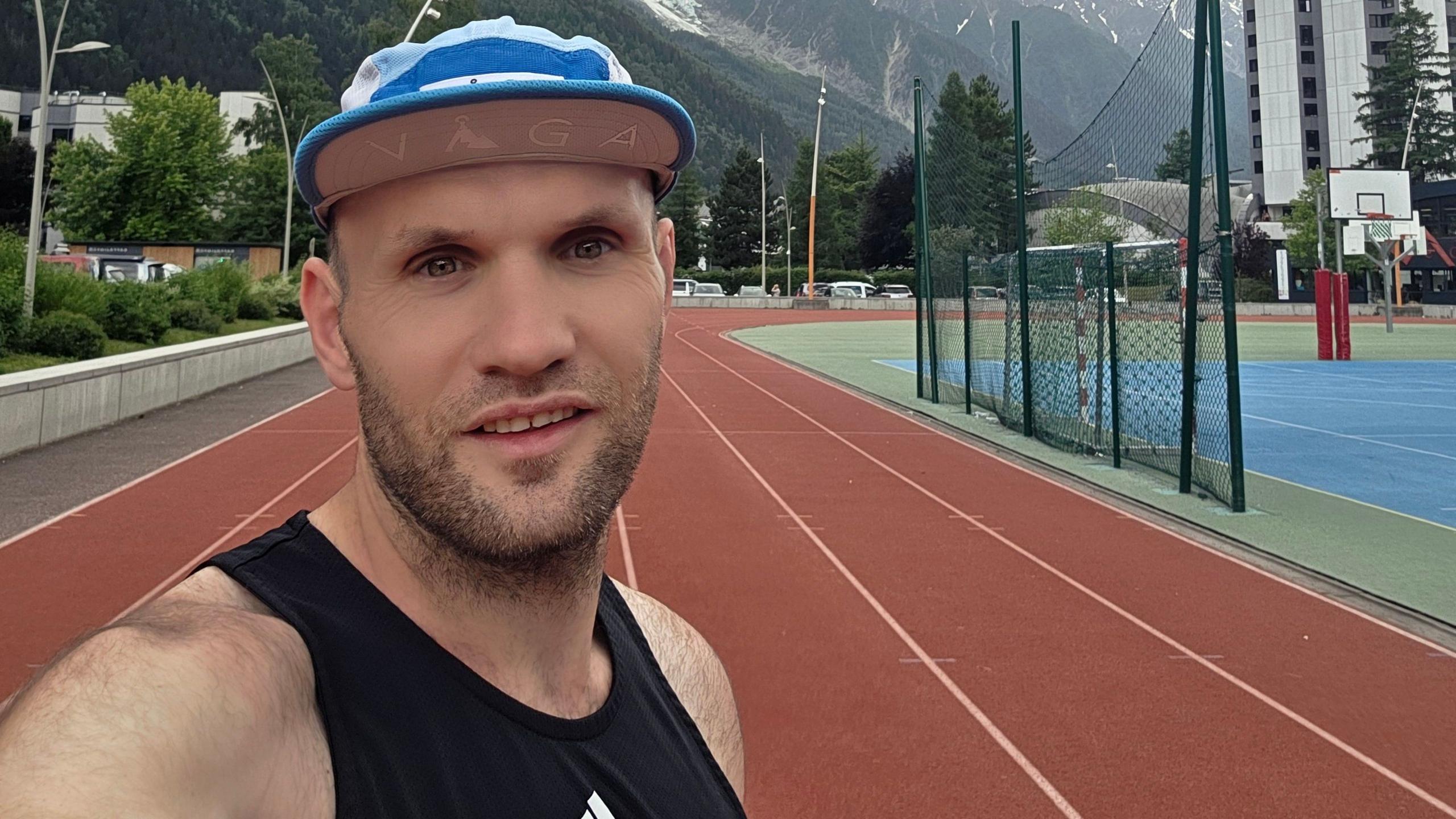 A man wearing a blue runners cap and a black training top. There is a running track and some mountains behind him.