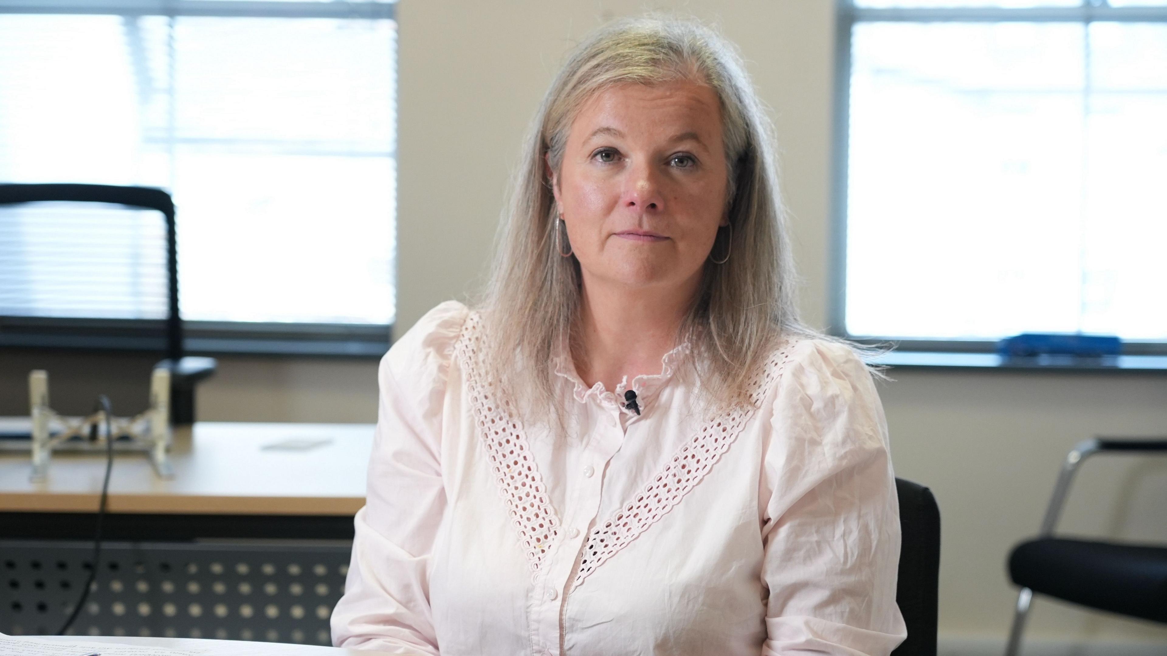 Lowri Griffiths is sitting behind a desk and is wearing a white shirt with a frilly neck. A small microphone is clipped to her collar and Lowri has shoulder length blonde/grey hair.
