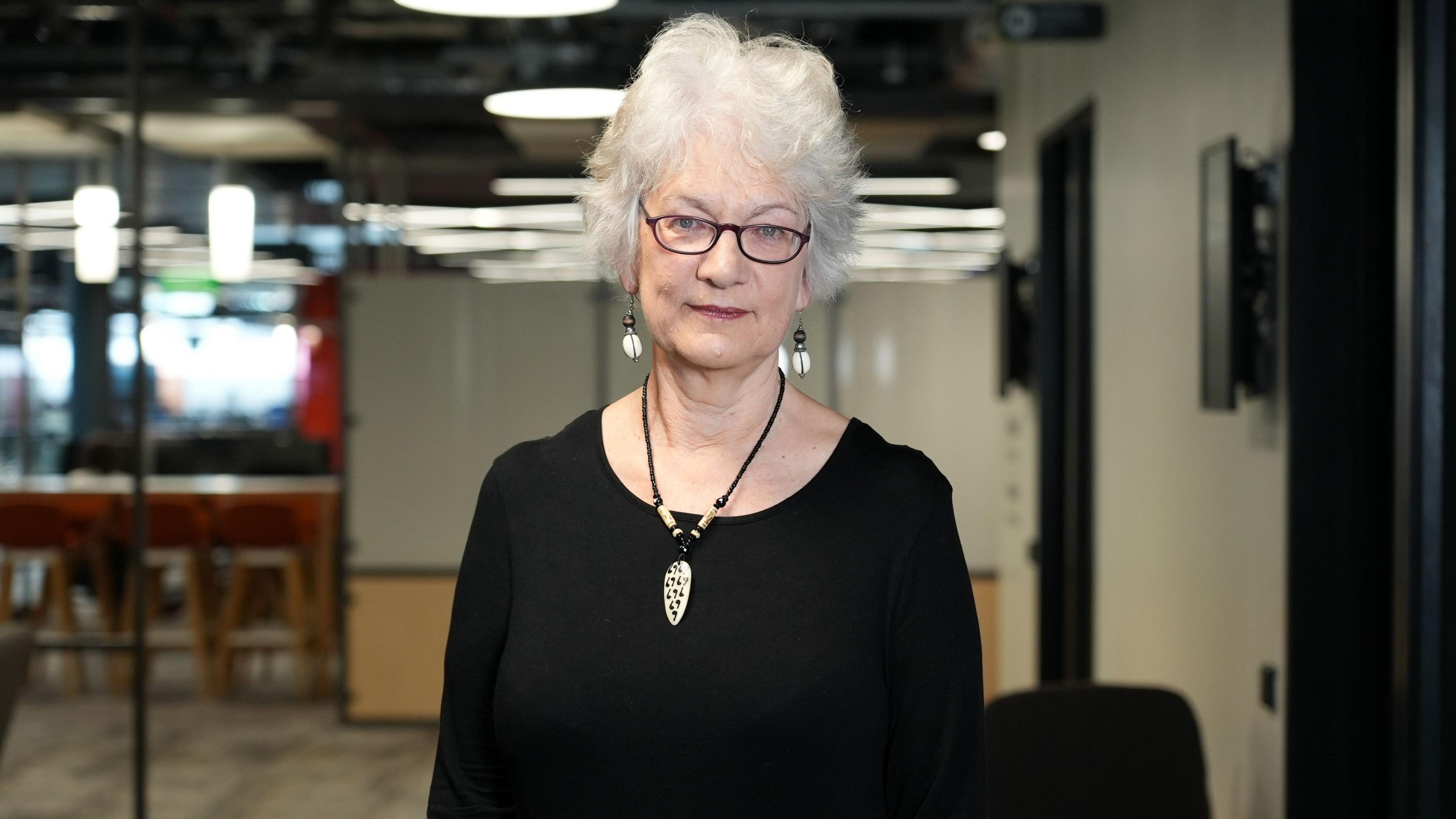 Dr Kathrin Thomas is inside an office building and is wearing a black, long-sleeved top, with matching earrings and a necklace. She has short white curly hair and is wearing dark, oval shaped glasses
