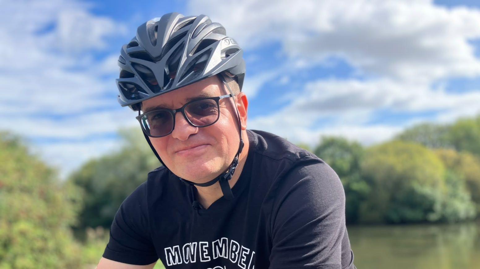 Ben Lidgey is wearing a grey cycling helmet and glasses. He is sat on a bicycle. Ben is wearing a black T-shirt with the visible text “MOVEMBER AMBASSADOR.” Trees and greenery are in the background with a partly cloudy sky.