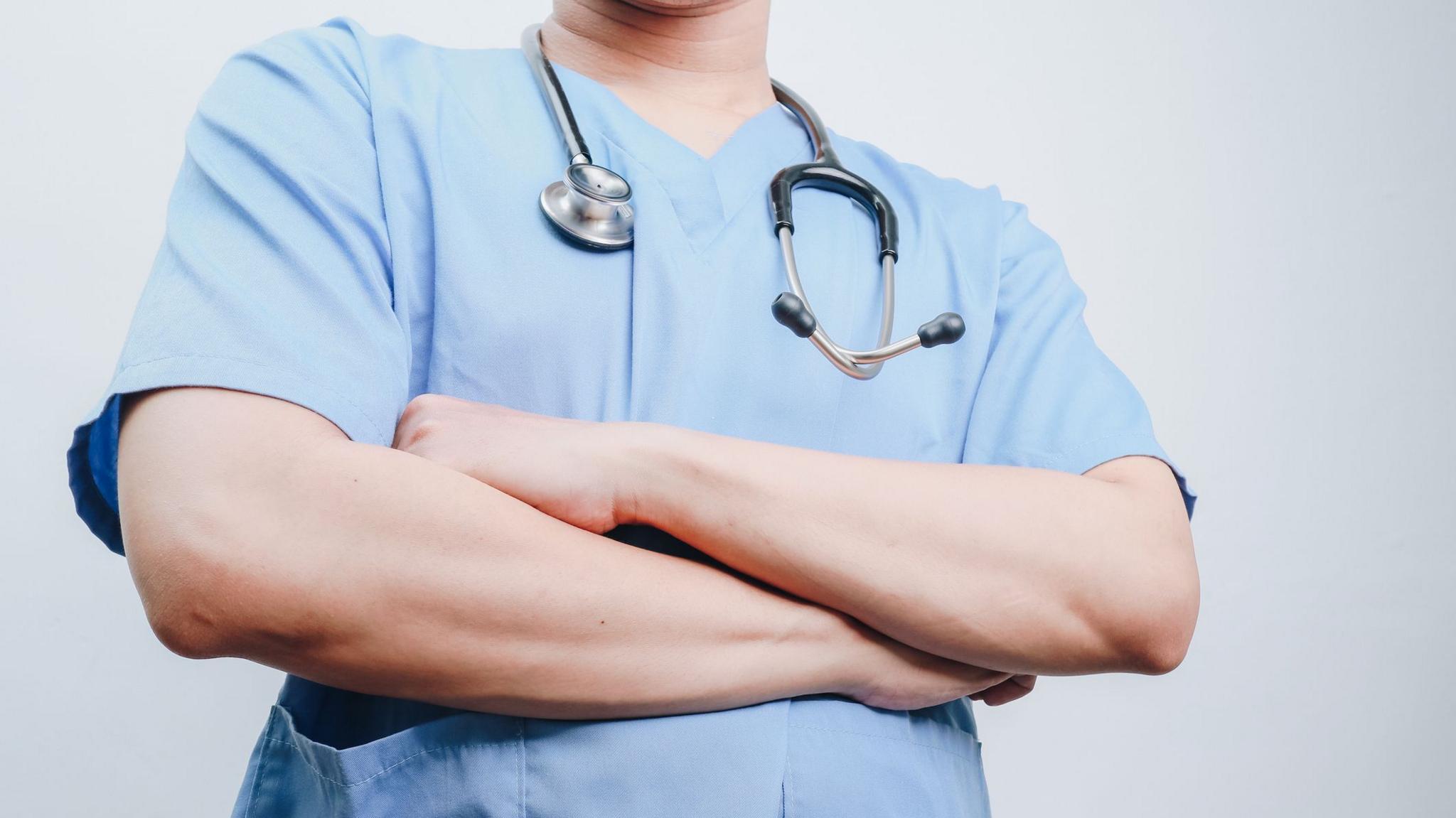 A person wearing medical scrubs has a stethoscope around their neck and is folding their arms.