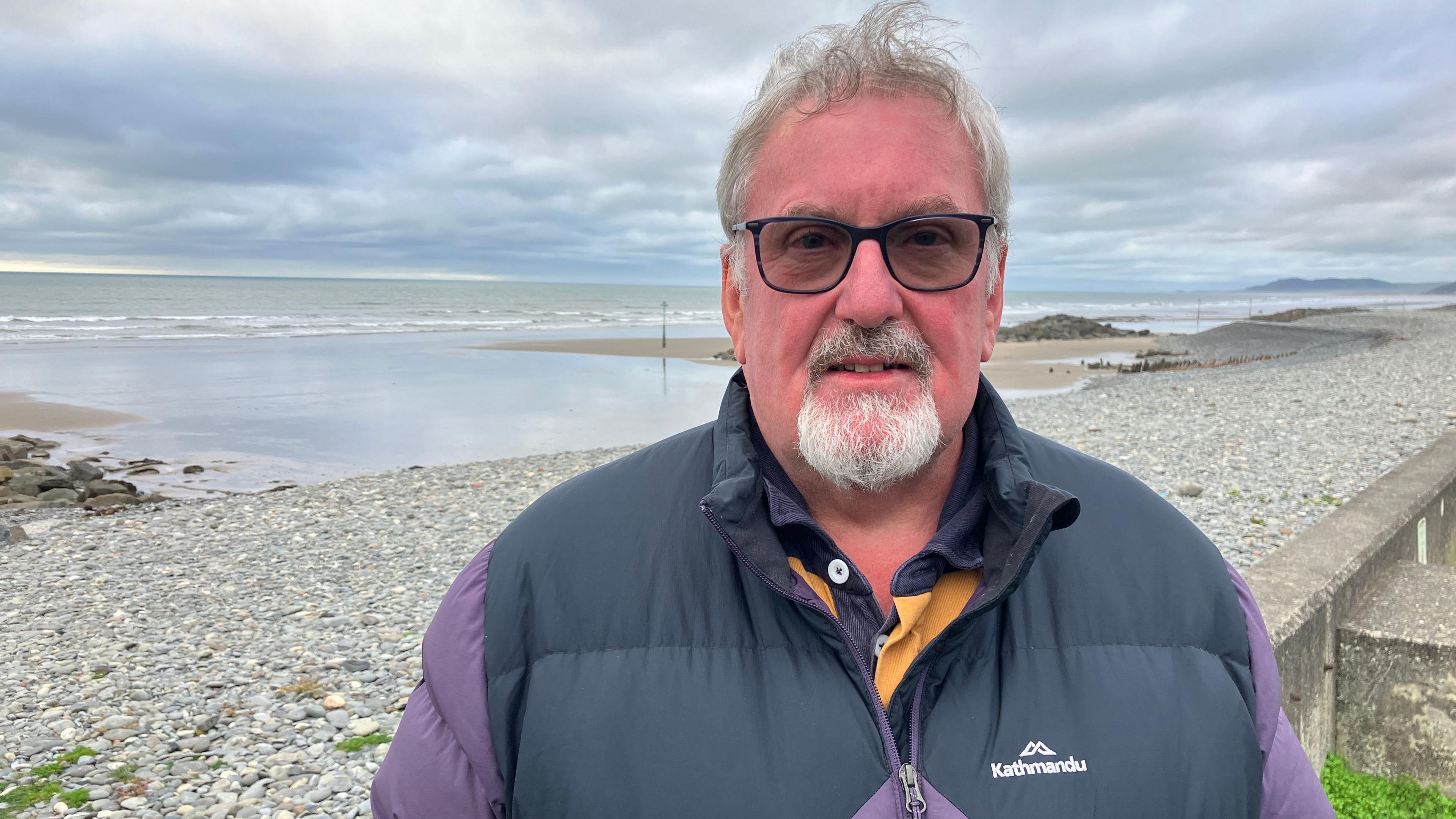 A man with grey hair and a goatee beard and moustache, wearing dark framed glasses and a navy blue and purple body warmer. He's looking straight at the camera and is standing in front of a pebble beach with the sea and sandbanks behind him in the middle distance