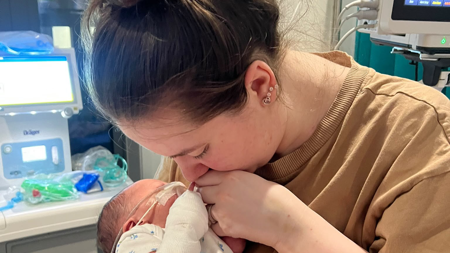 Sophieholding her newborn daughter Florence. Tubes can be seen attached to Florence's face. Her left arm is bandaged. Sophie has brown hair which is tied back. She wears a beige t-shirt. Her eyes are closed and she is not looking at the camera.