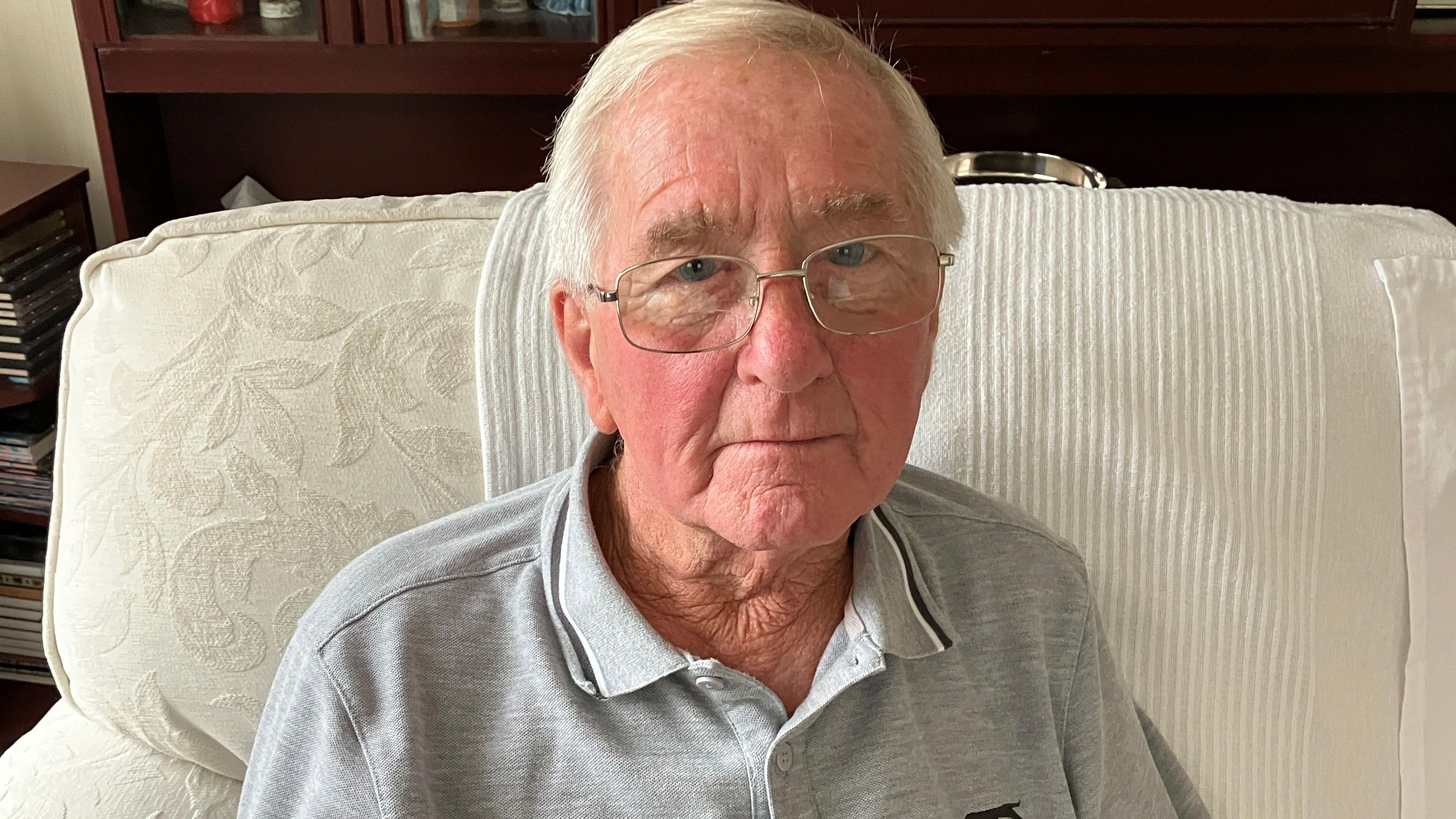 A photo of an older man - grey hair and glasses. He's sitting on a cream coloured sofa and is wearing a grey polo top