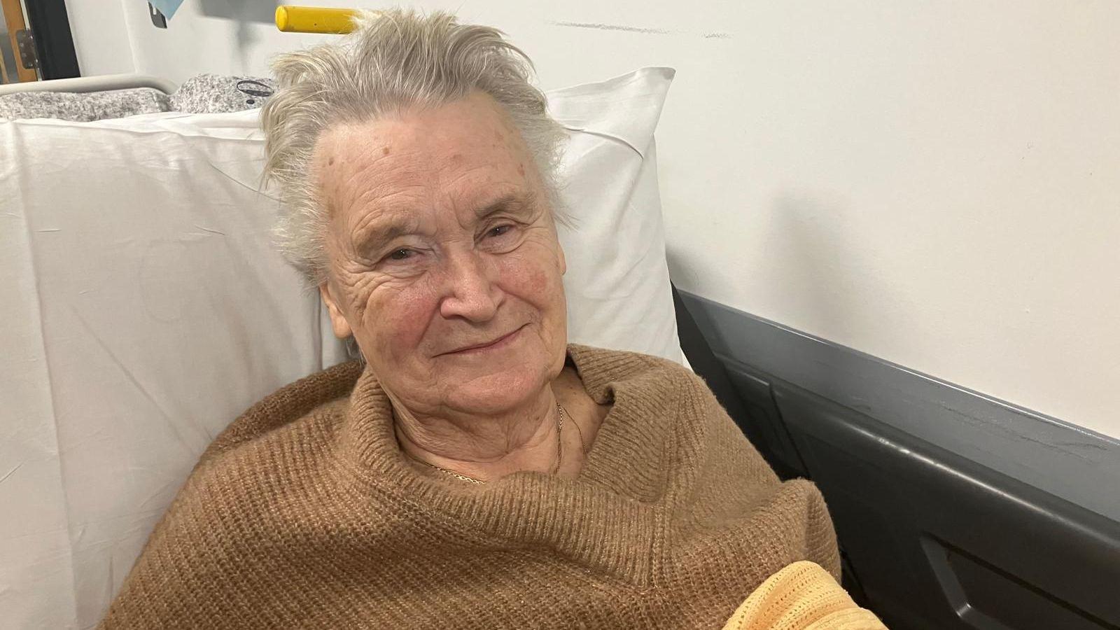 An woman with short silver hair is seen propped up on a hospital bed against a pillow. She wears a camel coloured wool jumper.