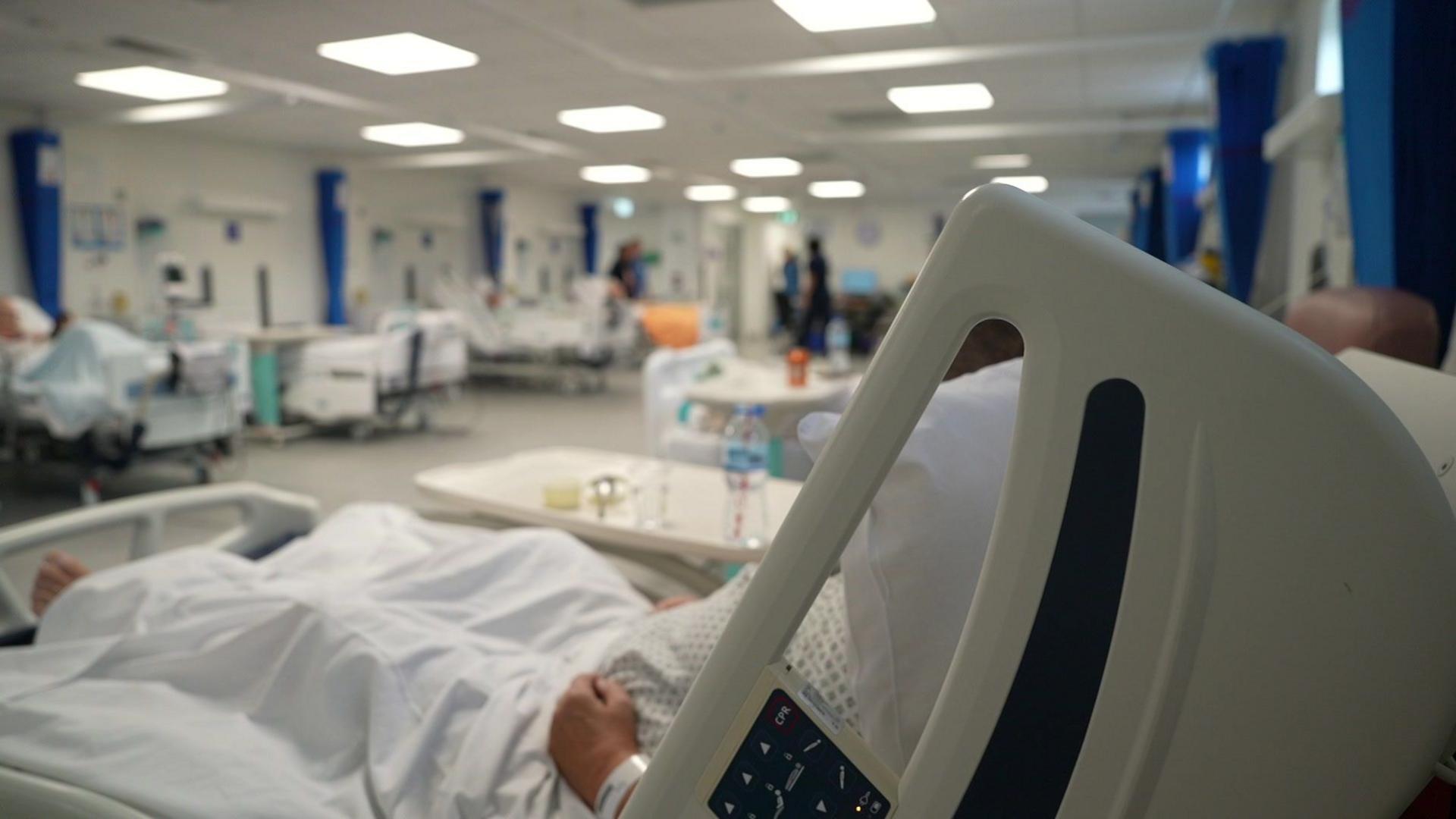 A hospital bed is seen in the foreground with a patient 's arm just visible with their head out of shot. A table holding a cup and a bottle of water can also be seen. In the background a number of other beds are seen out of focus.