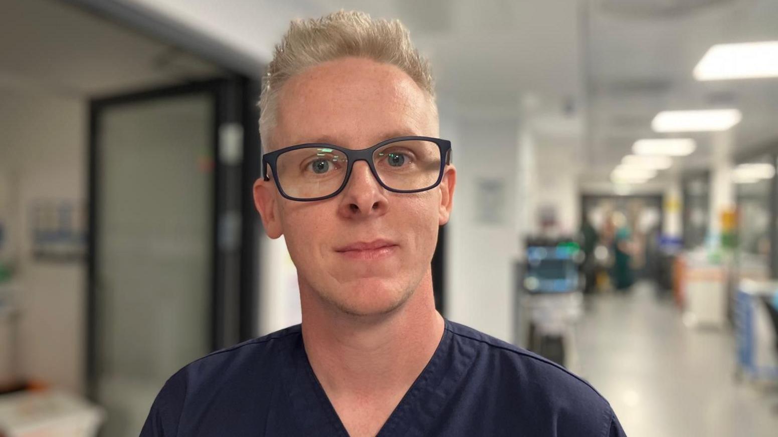 A young man smiles to camera. He has short cropped blonde hair and black rimmed oversize glasses. He wears dark navy v-neck scrubs.