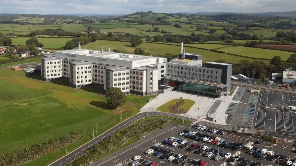 A large hospital building with car parks, and views across fields and mountains in the background.