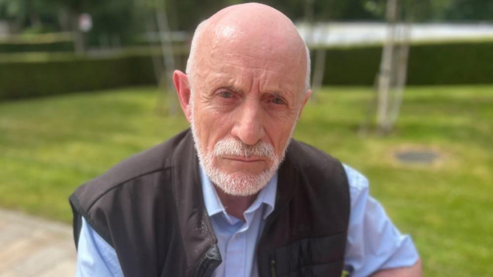 A man with a white beard looks into the camera. He is bald and is wearing a blue shirt and black gilet jacket.