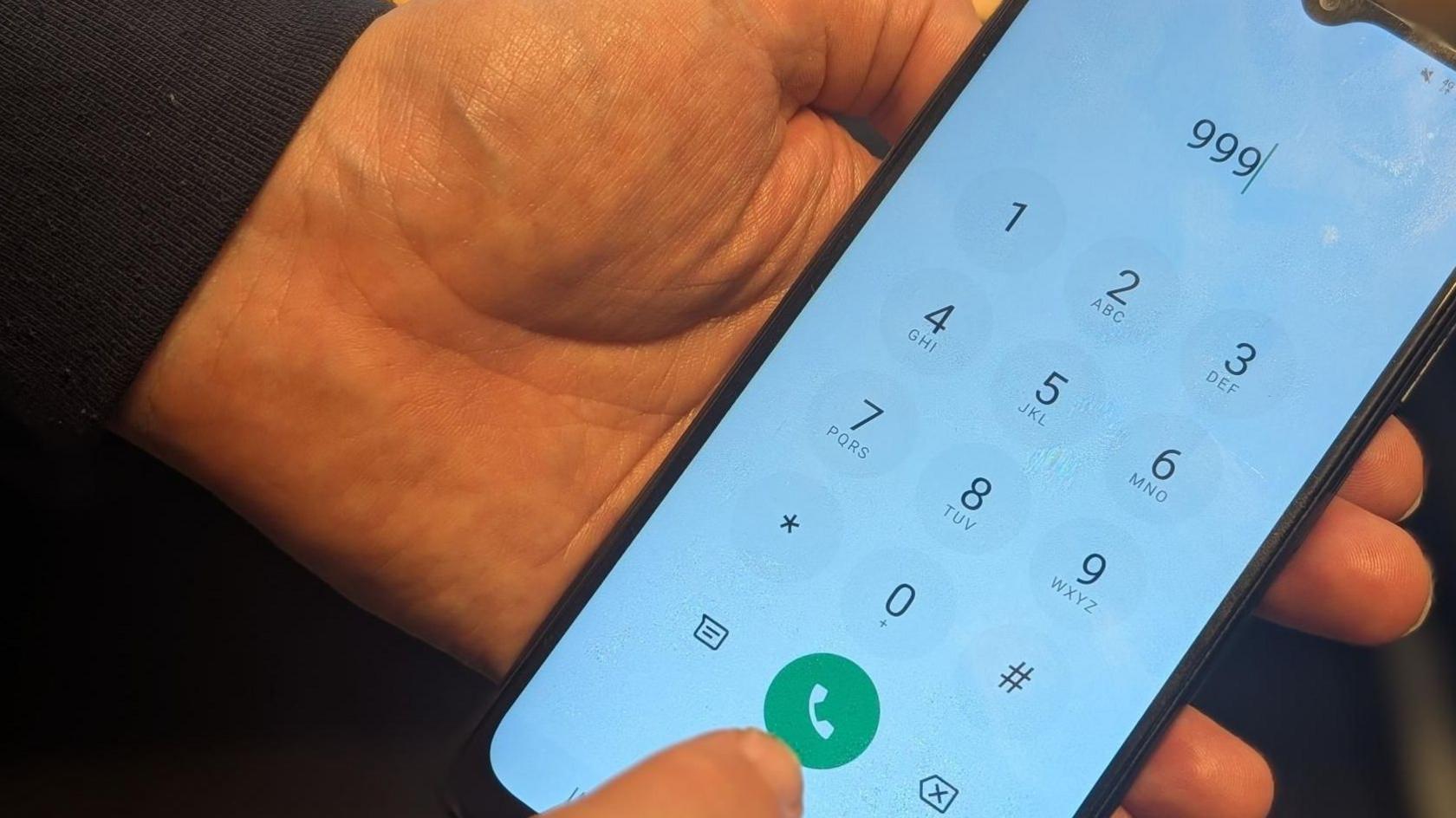 A close up image of a man's hand holding a mobile phone. He had dialled 999 and is about to press the green call button.