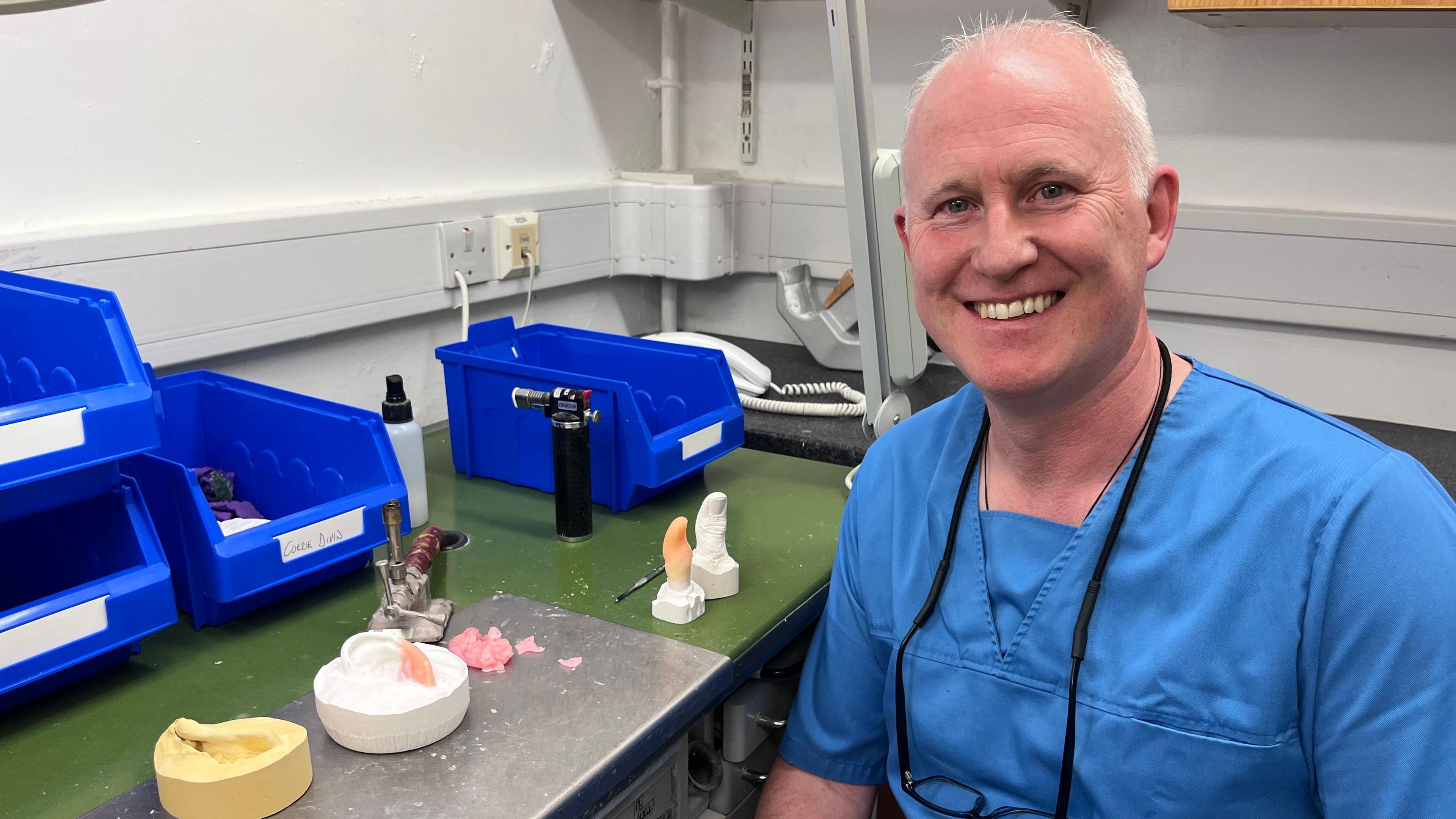 A man with cropped white hair, wearing blue scrubs in a hospital laboratory setting. He is sitting at a table with prosthetics on it. 