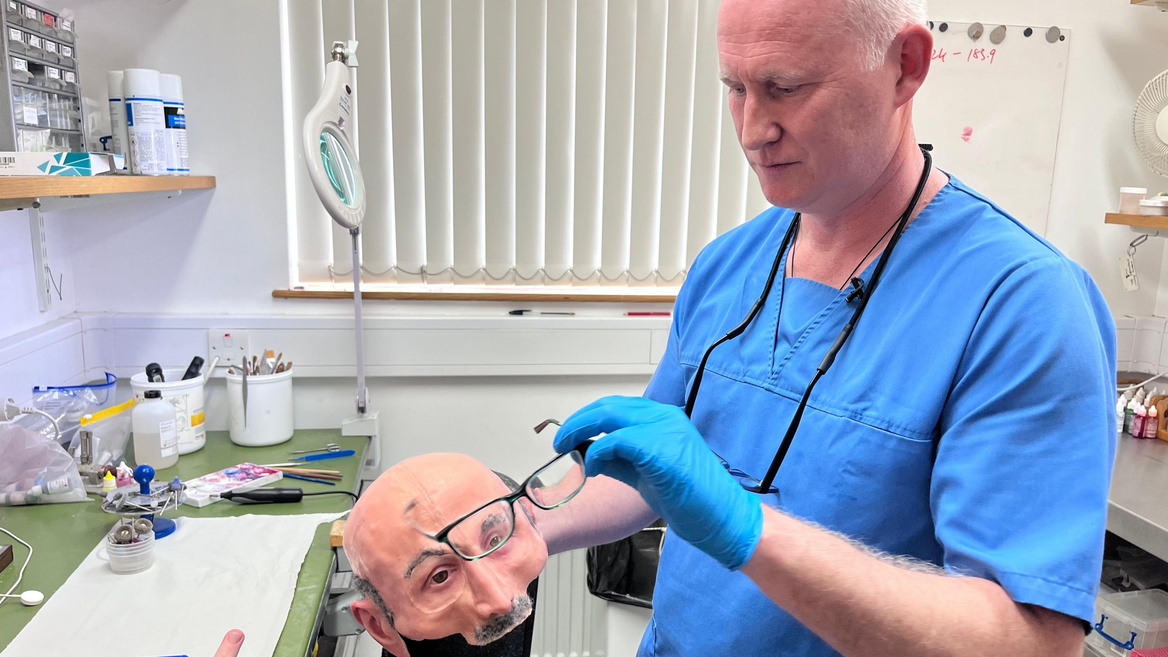 A doctor, with balding white hair, wearing blue scrubs. He is holding a prosthetic face and glasses. 