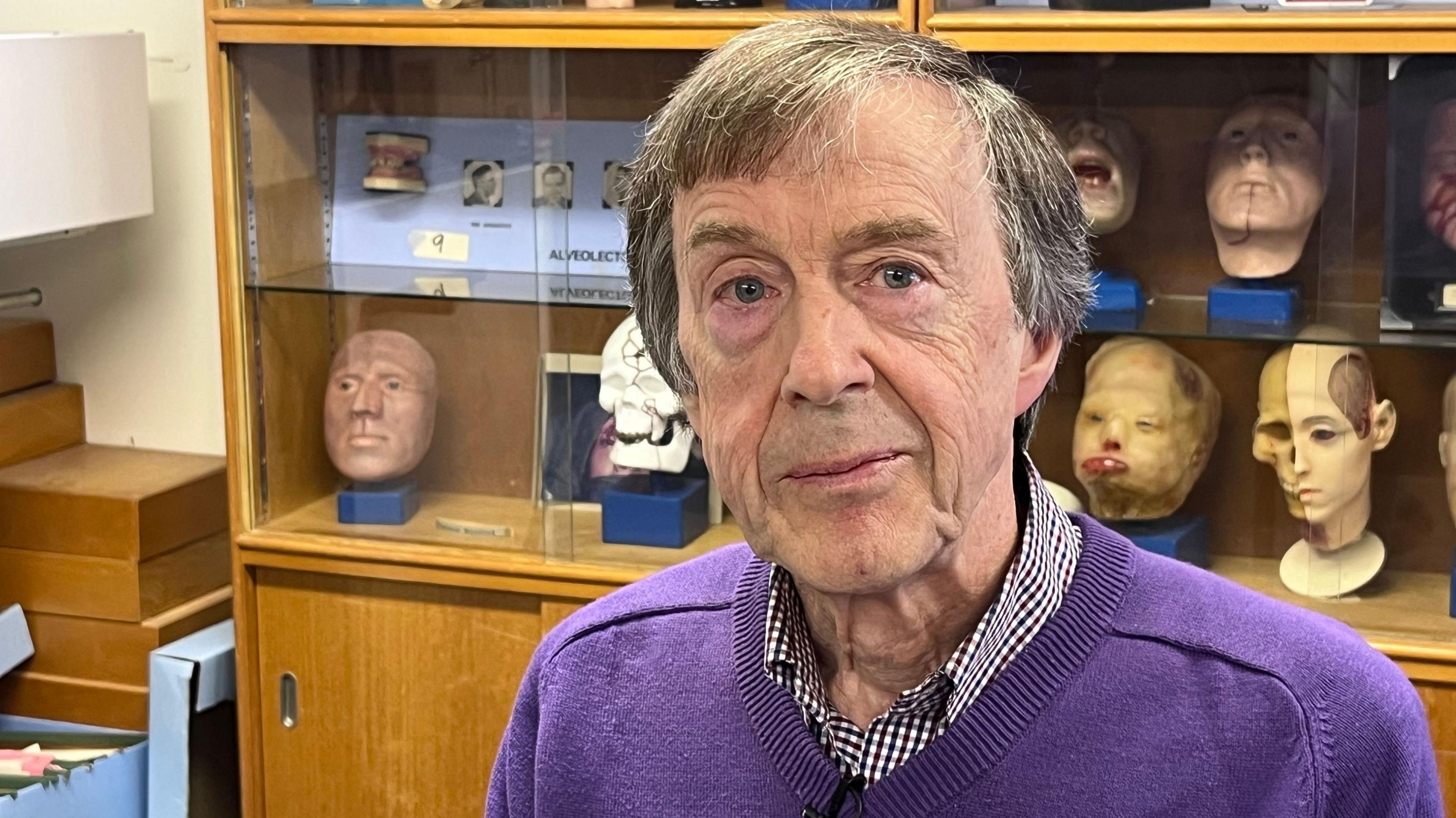 An older man, with short, dark grey hair, wearing a check shirt and a purple jumper. He is sitting in front of a cabinet with prosthetics inside. 