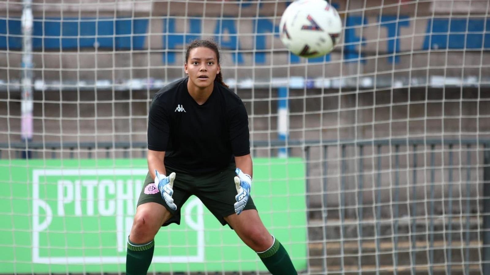 A young woman with dark hair tied back, wearing a black football shirt and shorts and padded white goalkeeper gloves is standing poised in the goal mouth. Her hands are out ready and her expression is completely fixed on the ball which is heading towards her.