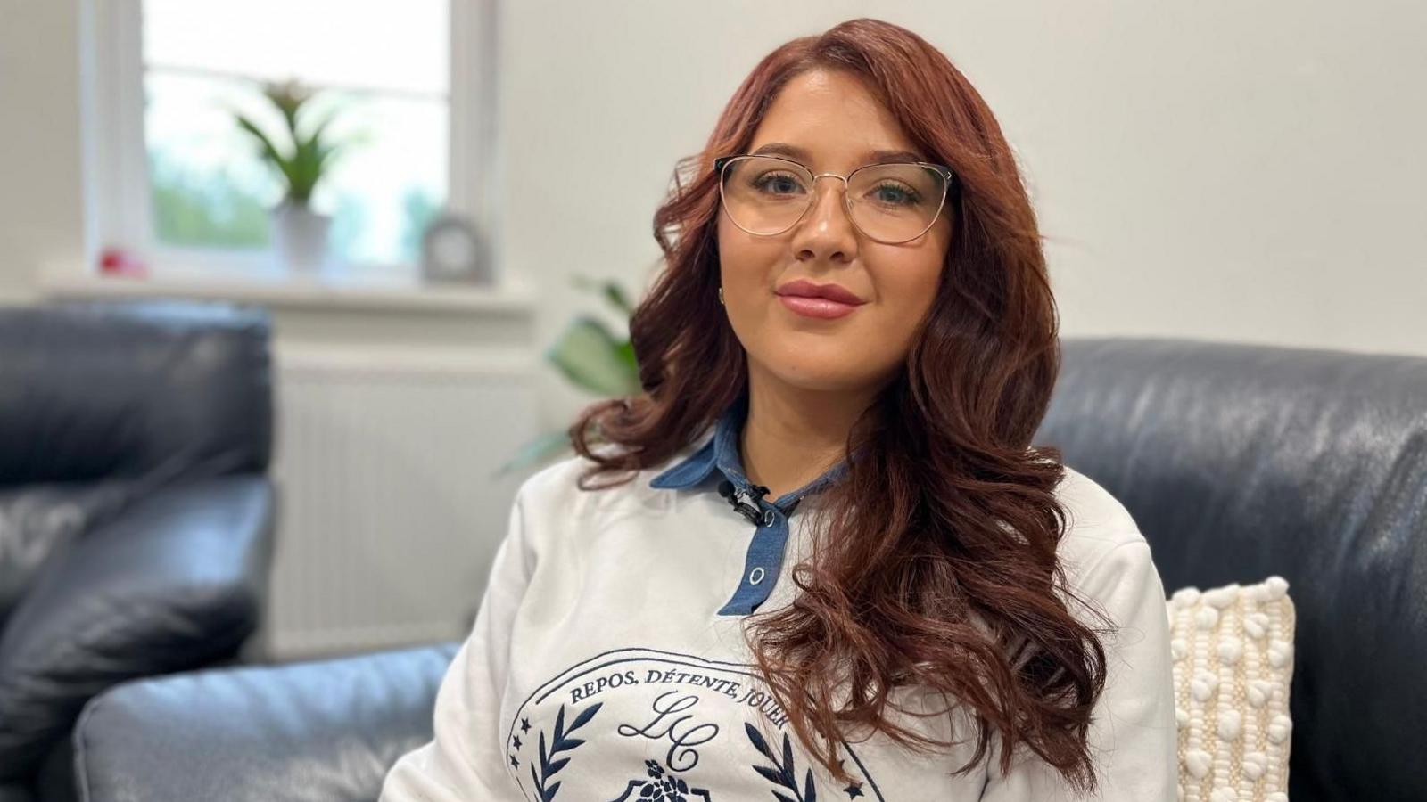Megan has long, wavy reddish brown hair. She is wearing glasses, a white and blue top, and is smiling at the camera. She is sat on a dark leather chair, with a window and white walls in the background