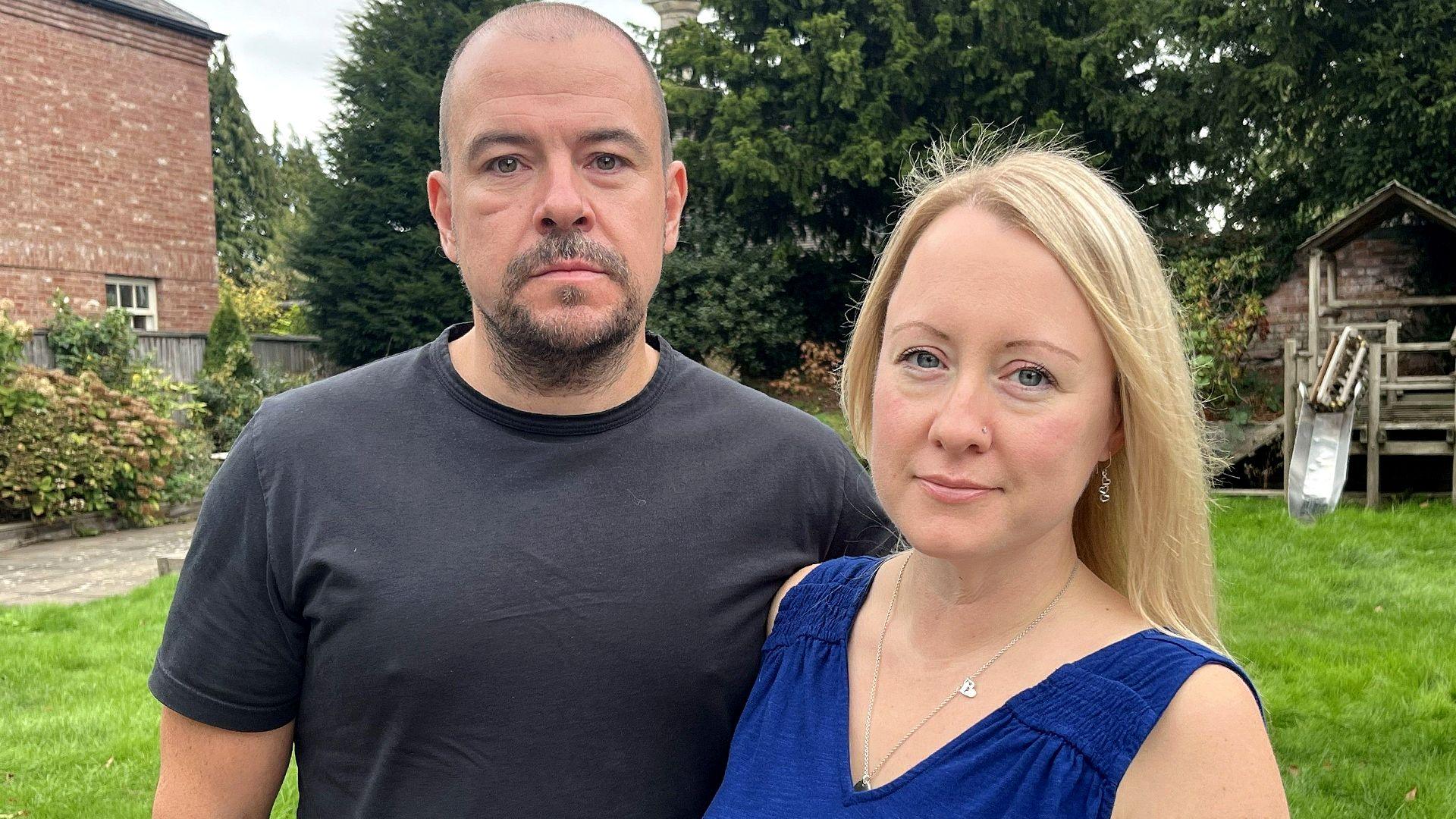 Neil Russell has a beard and is wearing a dark t-shirt. Next to him his wife Katie is wearing a blue sleeveless dress and has long blonde hair. They are pictured standing on grass in a garden in front of a brick building. 