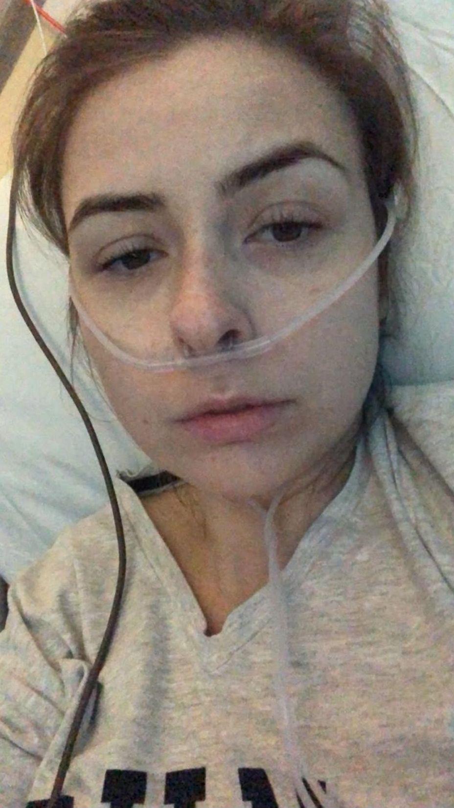 Kelly in a hospital bed, with a clear tube running out of her nose. Her hair is tied back and she wears a cream shirt. 
