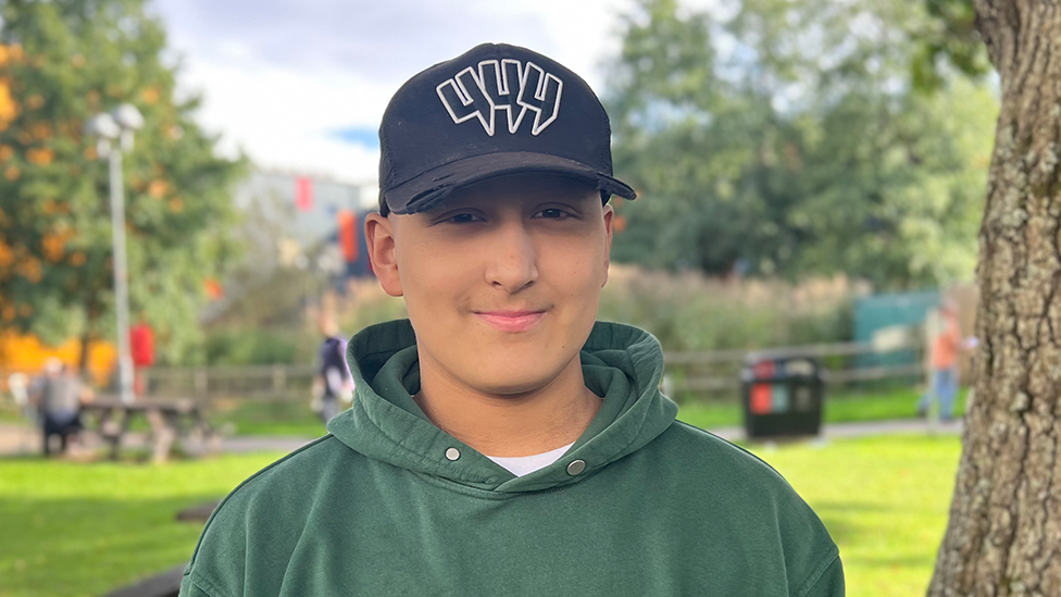Ethan looks at the camera and is wearing a dark green hoodie and a black baseball cap with a white logo. He is standing outdoors beside a tree. Grass, more trees, picnic benches and several people are visible out of focus in the background.