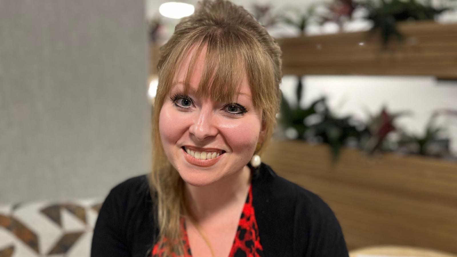 Delyth is wearing a red and black dress with a black cardigan. She is smiling at the camera and is sat with plants in the background. She has a fringe, with her brown hair tied back. 
