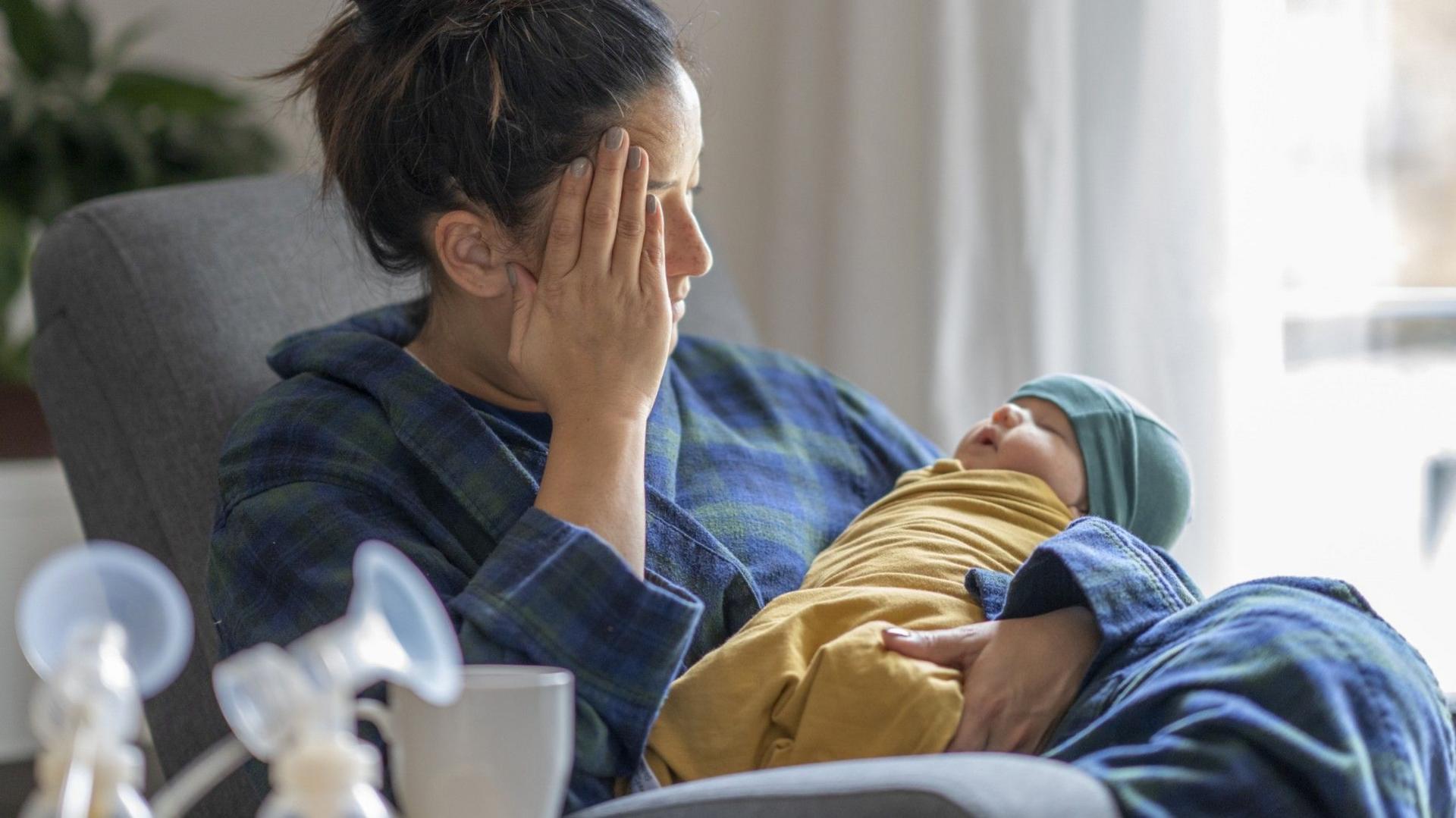 A stock image showing a mother wearing blue and green plaid pyjamas, sitting in a grey armchair holding her baby. The baby is asleep, swaddled in a yellow cloth wearing a green hat. The woman has her dark hair tied up in a messy bun and is covering her face with her hand, looking tired and exasperated. In the blurred foreground there are two breast pumps visible, and in the background there is a bright window with a white curtain.