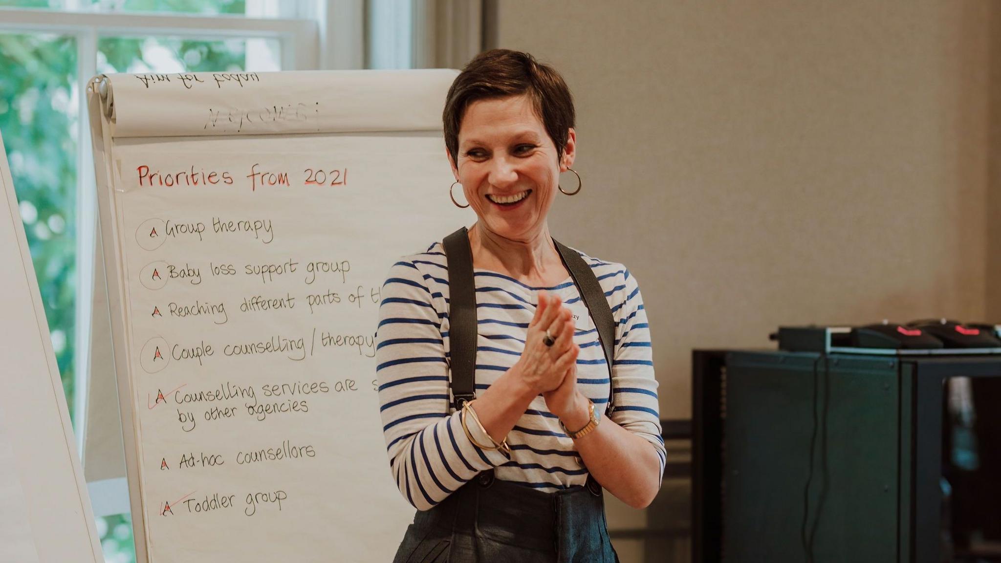 Suzy Cristinacce is standing next to a flip board with writing on it. She is clapping her hands together and laughing.