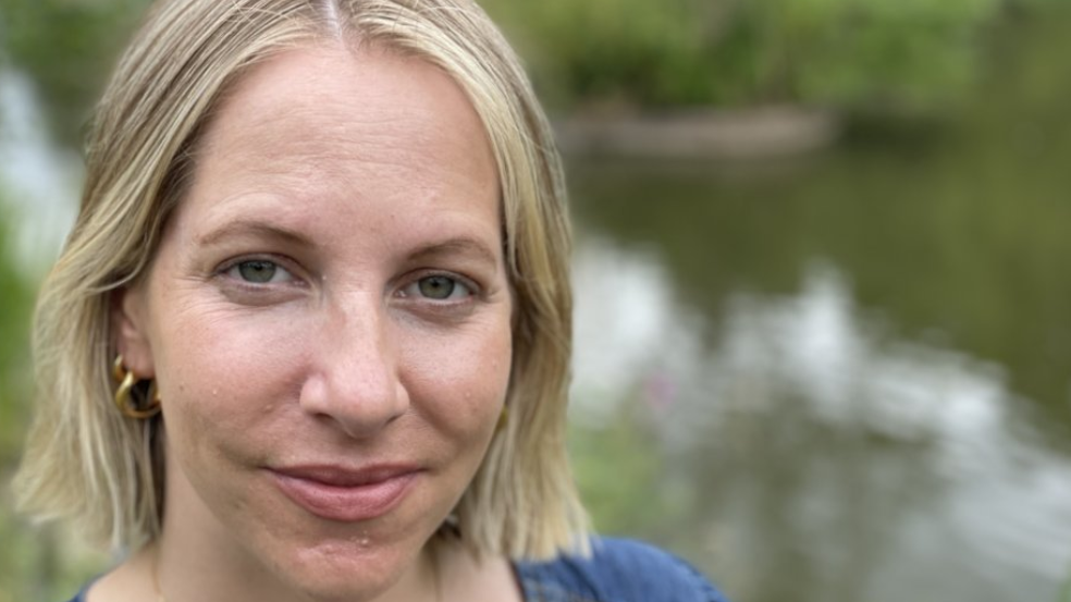A headshot of a woman with a blonde bob, smiling at the camera. She is standing in front of a river, which is blurred out in the background. She has gold hoop earrings in and is wearing a blue top.