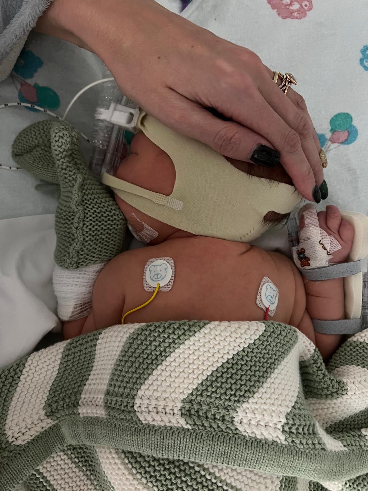 Newborn Phoebe lying on a hospital cot, partially covered with a green-and-white knitted blanket. The baby is wearing medical monitoring leads, a respiratory support mask secured with a soft headband, and protective mittens. An adult hand rests gently on the baby's head. The hospital bedding has a subtle balloon pattern in the background.