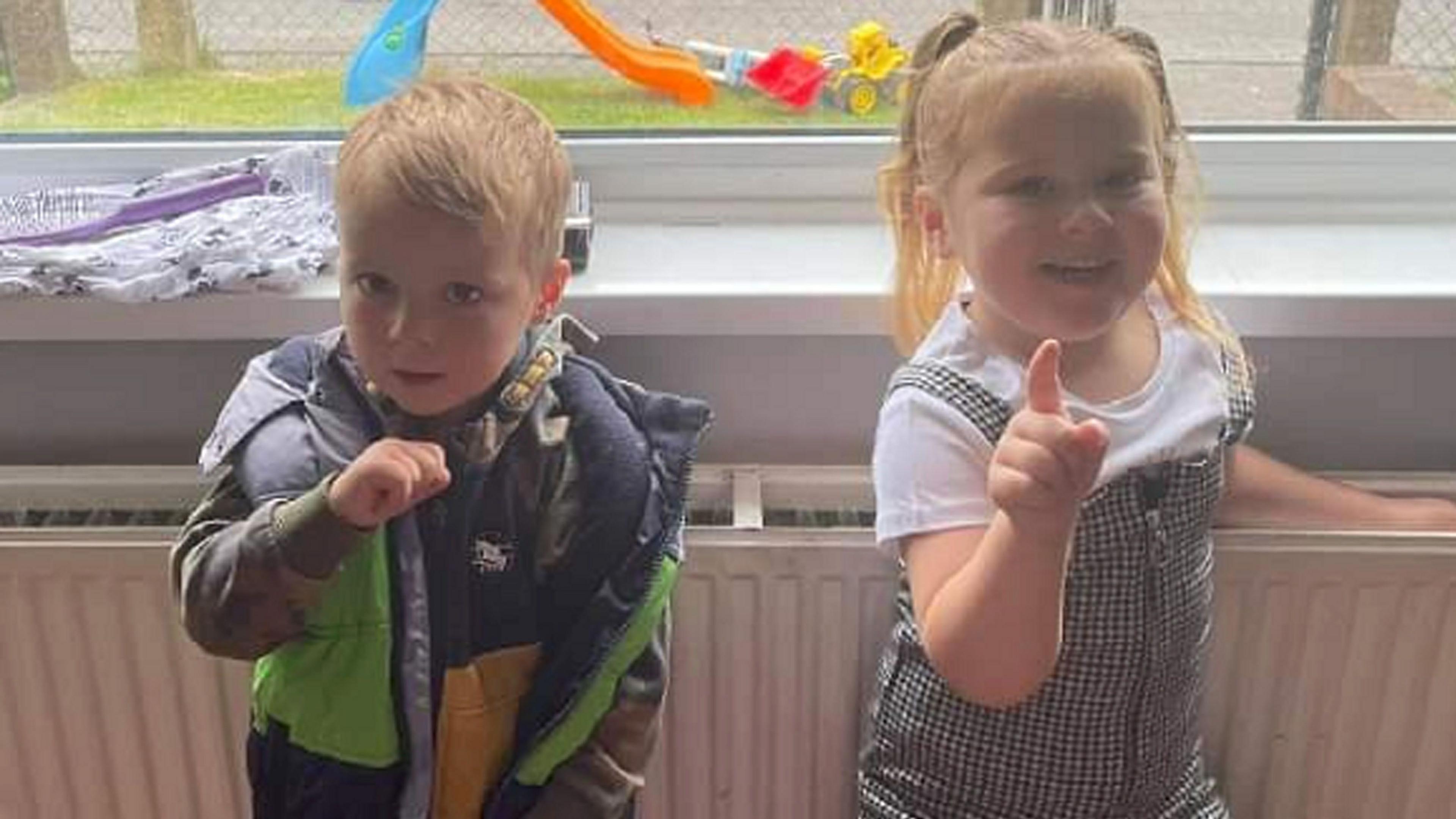 Two children looking at the camera. The young blond boy wears a green and black coat with his hand in the air. A young girl with her blonde hair in ponytails and wearing a black chequered dress is pointing. They are both standing against a white radiator. 