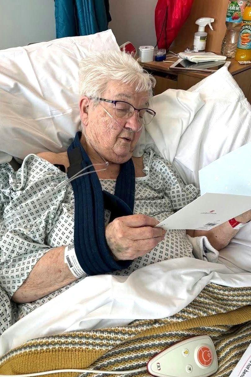 Margaret Owen is sat upright in bed. She is holding a card in her hands and looks down to read it. A navy sling can be seen on her left arm. She has short silver hair and wears a white hospital gown and reading glasses. A control for the bed can be seen on the bed.