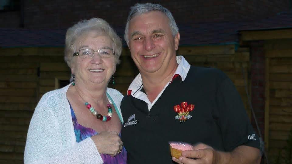 Graham Anthony (right) pictured with his mum Margaret. He has silver short hair, wears a navy welsh rugby jersey and smiles at the camera. Margaret is wearing a purple vest top with a white cardigan and holds her left hand to her chest. She has blue eyes, wears glasses, and smiles at the camera.