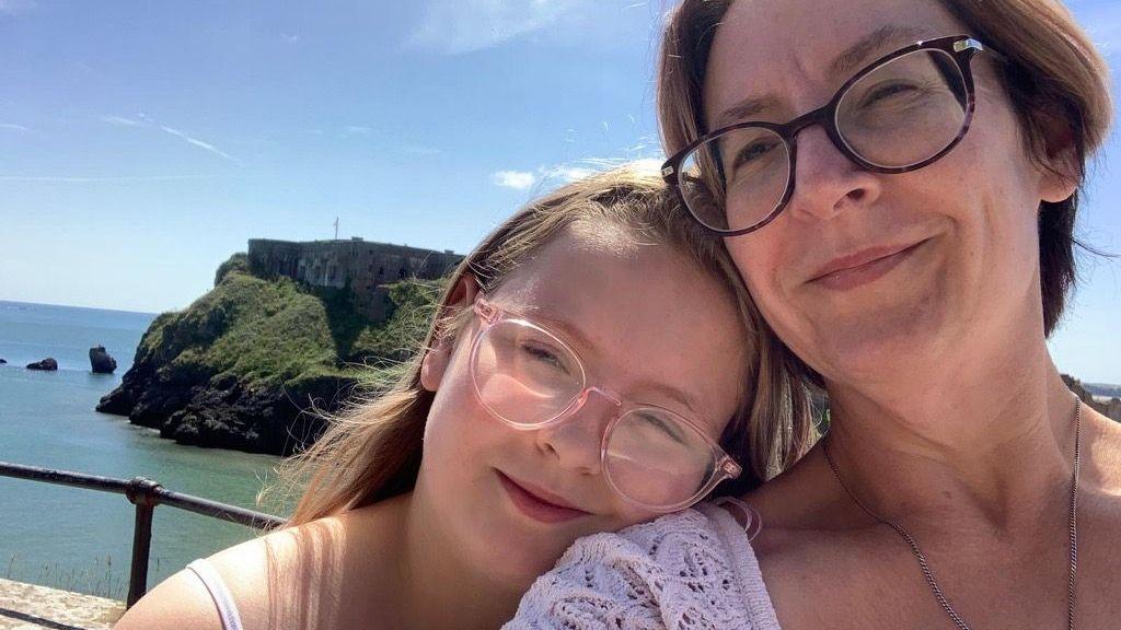 A young girls wearing glasses with blonde hair rests her chin on the shoulder of a woman with brown hair and glasses. An island surrounded by water is seen behind them.