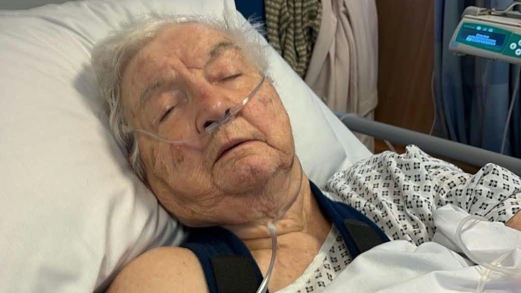 Margaret Owen is led down in a hospital bed, with a breathing tube under her nose. She has short silver hair and her eyes are shut.